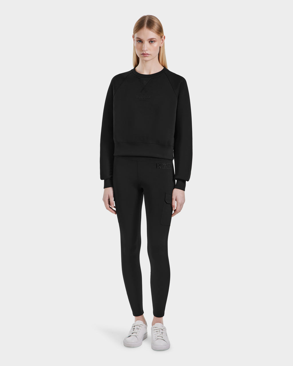 A woman with long blonde hair and a neutral expression wears the SIEN - 8525090 Black activewear set by Rudsak—matching sweatshirt and high-rise leggings with white sneakers—standing against a plain white background.
