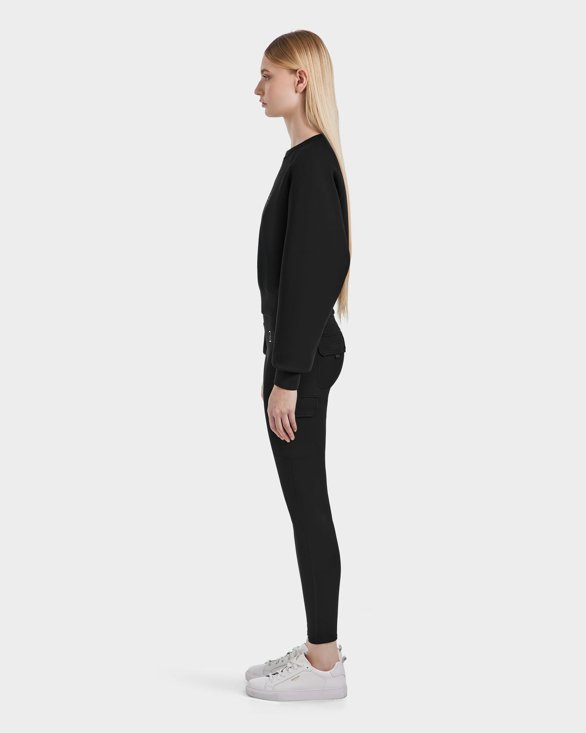 A person with long blond hair stands in profile facing left, wearing the Rudsak SIEN - 8525090 Black long-sleeve top, high-rise leggings, and white sneakers against a plain light gray background.
