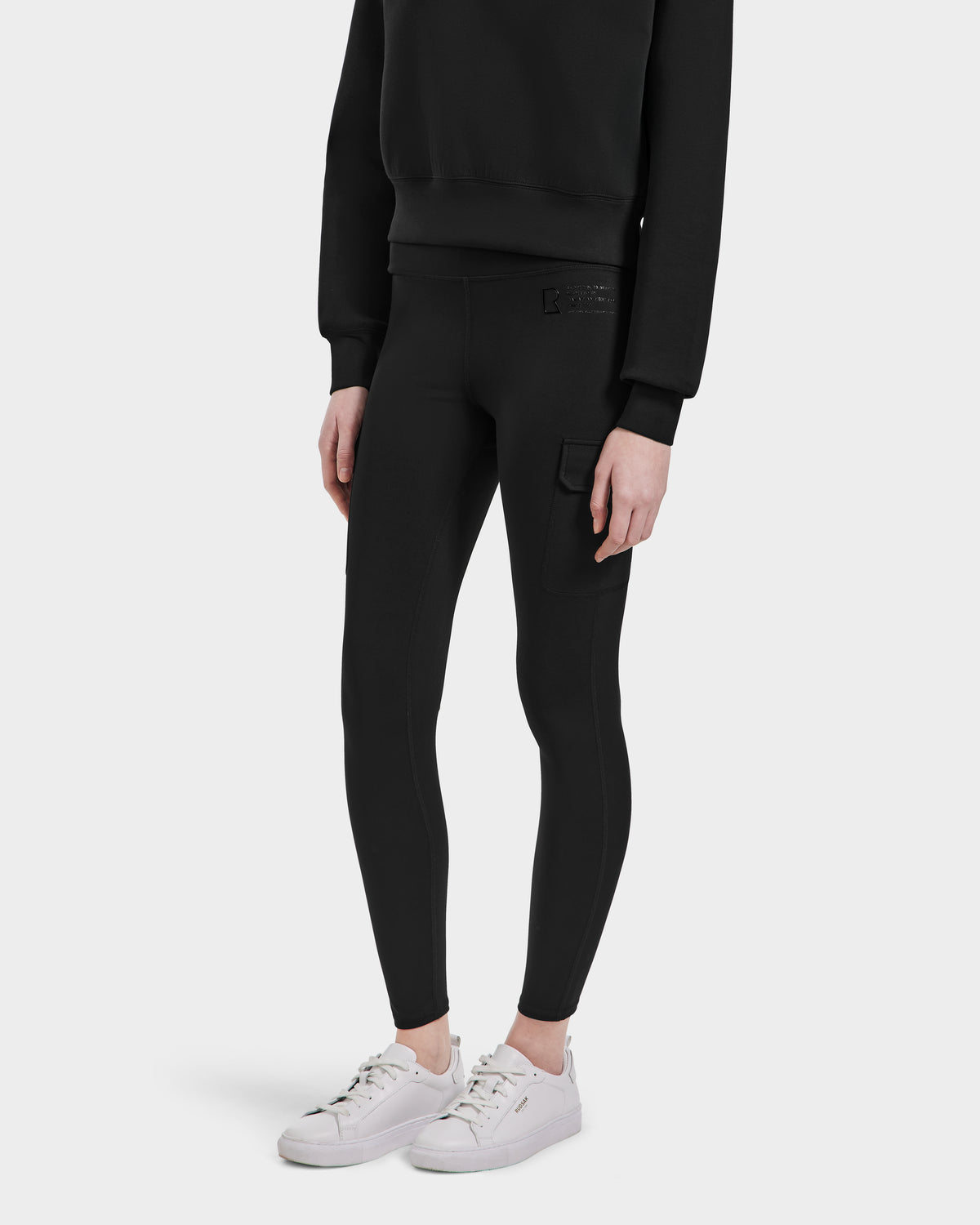 Wearing Rudsak’s SIEN - 8525090 Black sweatshirt and high-rise leggings with a side pocket, paired with white sneakers, an individual stands against a plain white background. Their face is not shown.