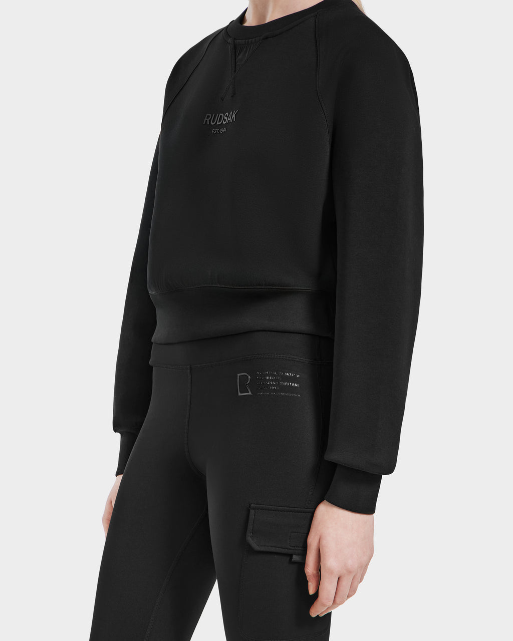 A person wears the Rudsak SIEN - 8525090 Black sweatshirt with RUDSAK EST 1994 on the chest and high-rise cargo pocket leggings, standing against a white background—essential activewear for everyday comfort.