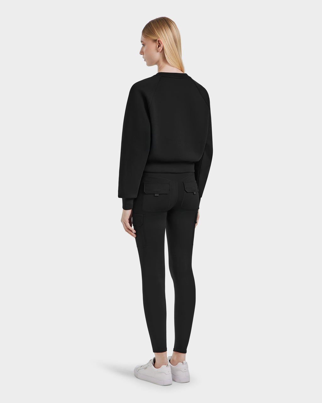 A woman with straight blonde hair stands facing away, wearing the Rudsak SIEN - 8525090 Black long-sleeve sweatshirt against a plain light gray background.