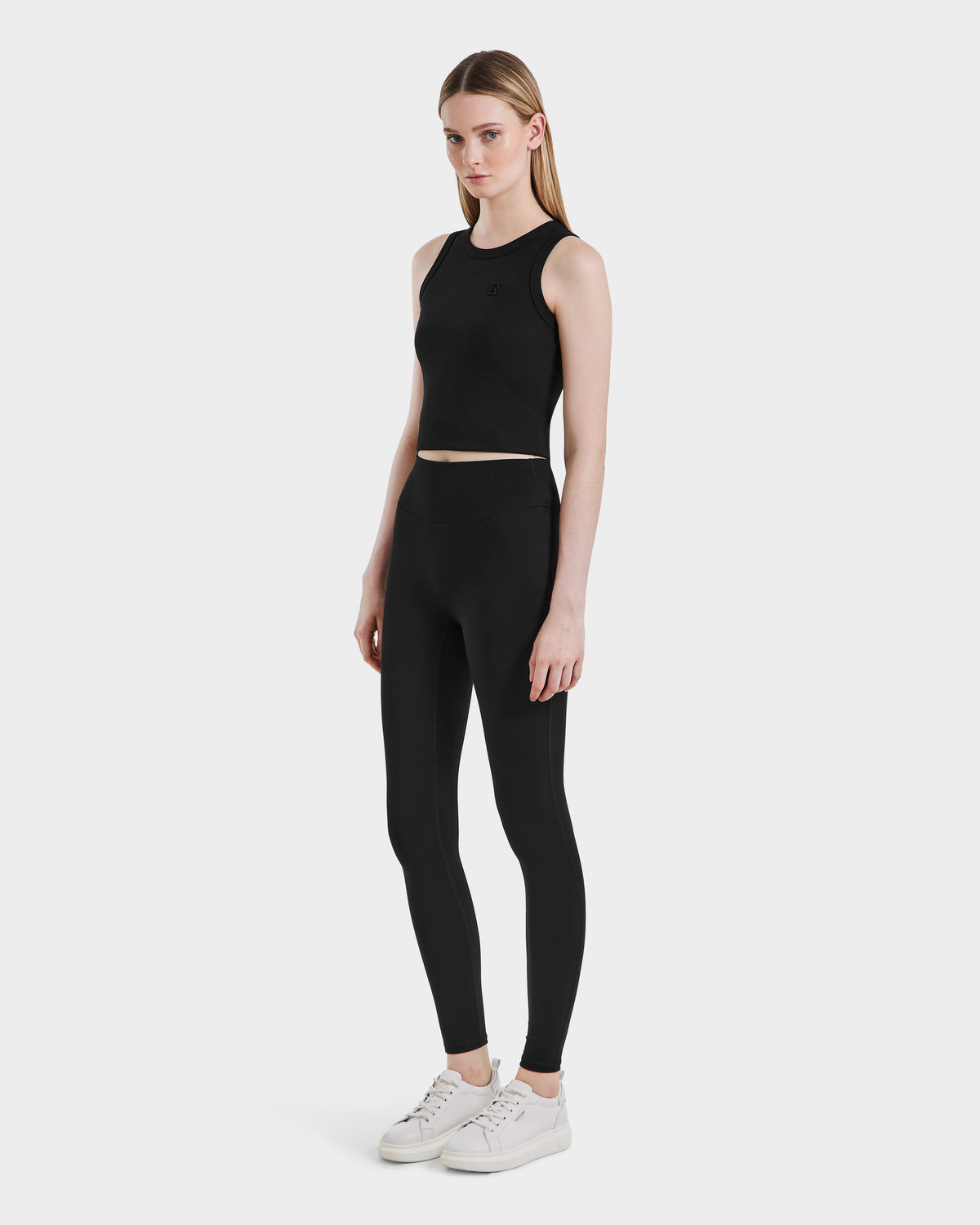 A woman with straight, long hair and a neutral expression stands against a plain background wearing Rudsak SONOMA - 8525071 Black high-performance leggings, a black sleeveless crop top, and white sneakers.