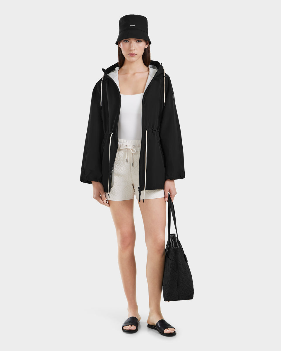 A woman stands against a plain white background, wearing the Rudsak SOPHIE - 8125009 Black bucket hat paired with a mid-length raincoat, white tank top, beige drawstring shorts, black sandals, and holding a black tote bag.