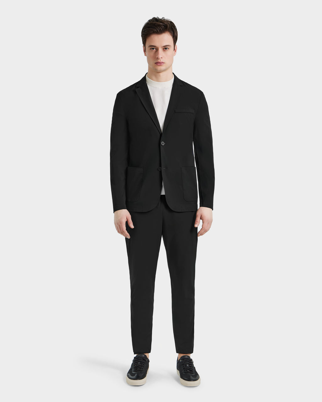 A man stands facing forward in the Rudsak SPENCER - 6525024 Black blazer and pants, paired with a white crew-neck tee and black sneakers—modern essentials—against a plain white background.