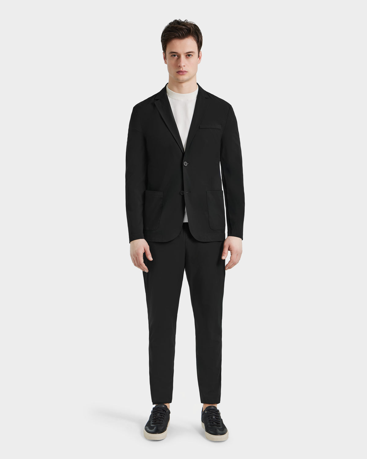 A man stands facing forward in the Rudsak SPENCER - 6525024 Black blazer and pants, paired with a white crew-neck tee and black sneakers—modern essentials—against a plain white background.