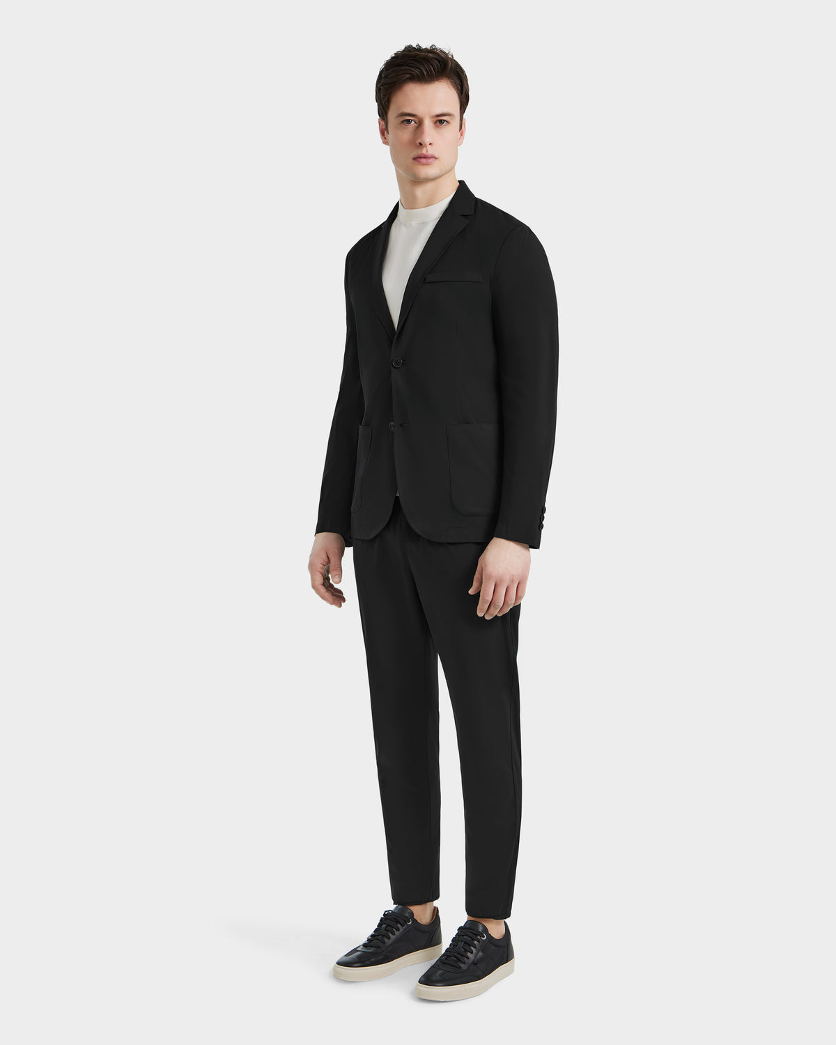 A man faces forward, styled in the SPENCER - 6525024 Black blazer by Rudsak over a white turtleneck and black sneakers. The minimalist light background brings out the sharp, modern contrast of his outfit.