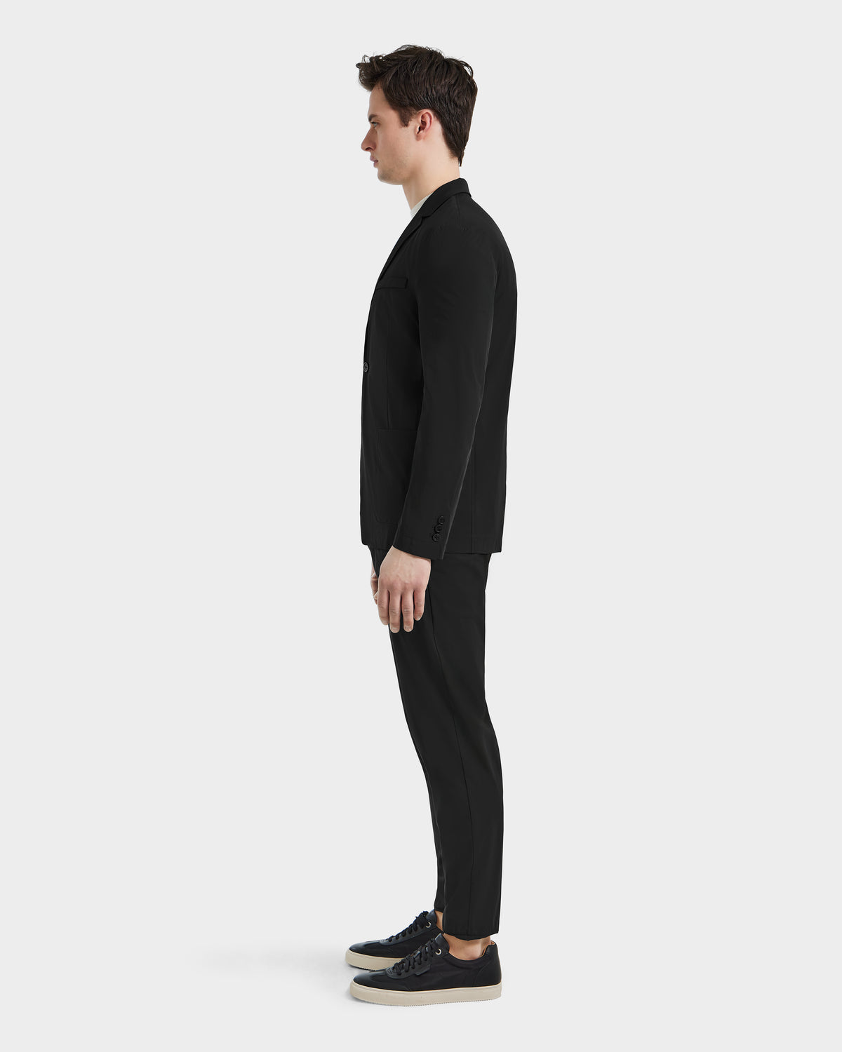 A man in the Rudsak SPENCER - 6525024 Black blazer and black suit with black sneakers stands in profile against a plain white background, highlighting a modern wardrobe.