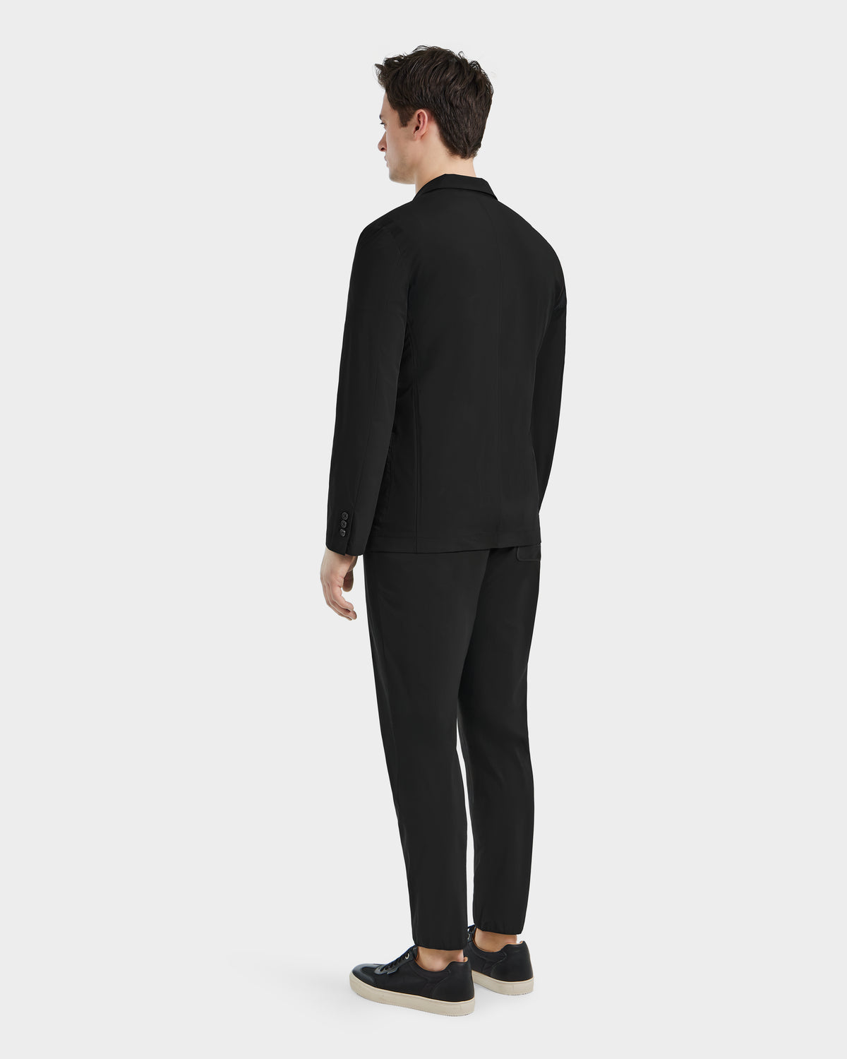 A man faces away from the camera, wearing a refined blazer and Rudsaks SPENCER - 6525024 Black sneakers with white soles—modern essentials—posed against a plain light background.