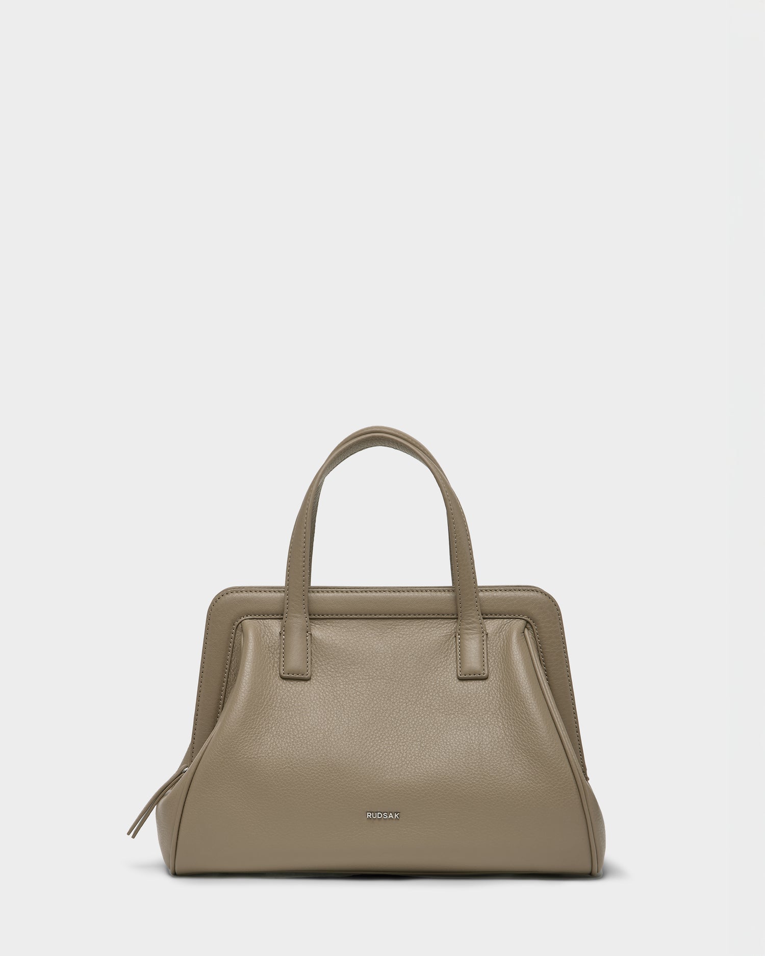 The Rudsak TAULLA - 8325002 Almond is a women’s pebble leather satchel in taupe with two handles, a structured rectangular design, metallic brand lettering on the front, and a spacious interior, shown against a plain white background.