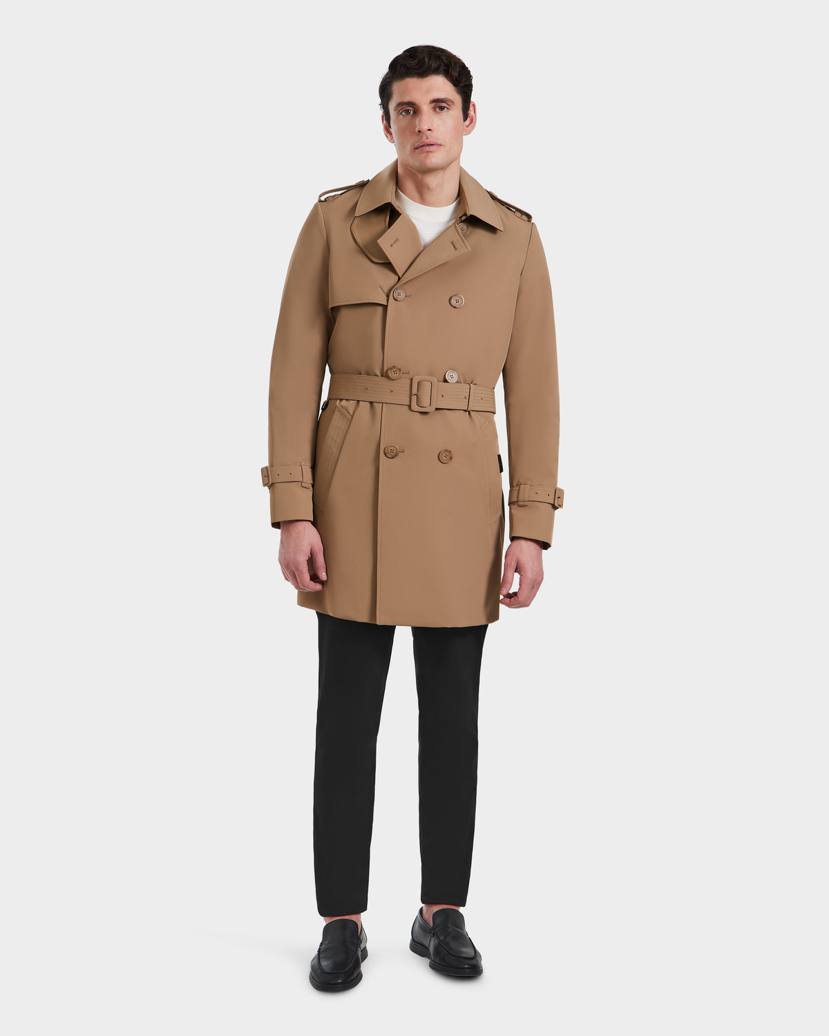 A man wearing the Rudsak TIM - 6124025 Camel trench coat with a belt, black pants, and black shoes stands against a plain white background, facing forward with a neutral expression.