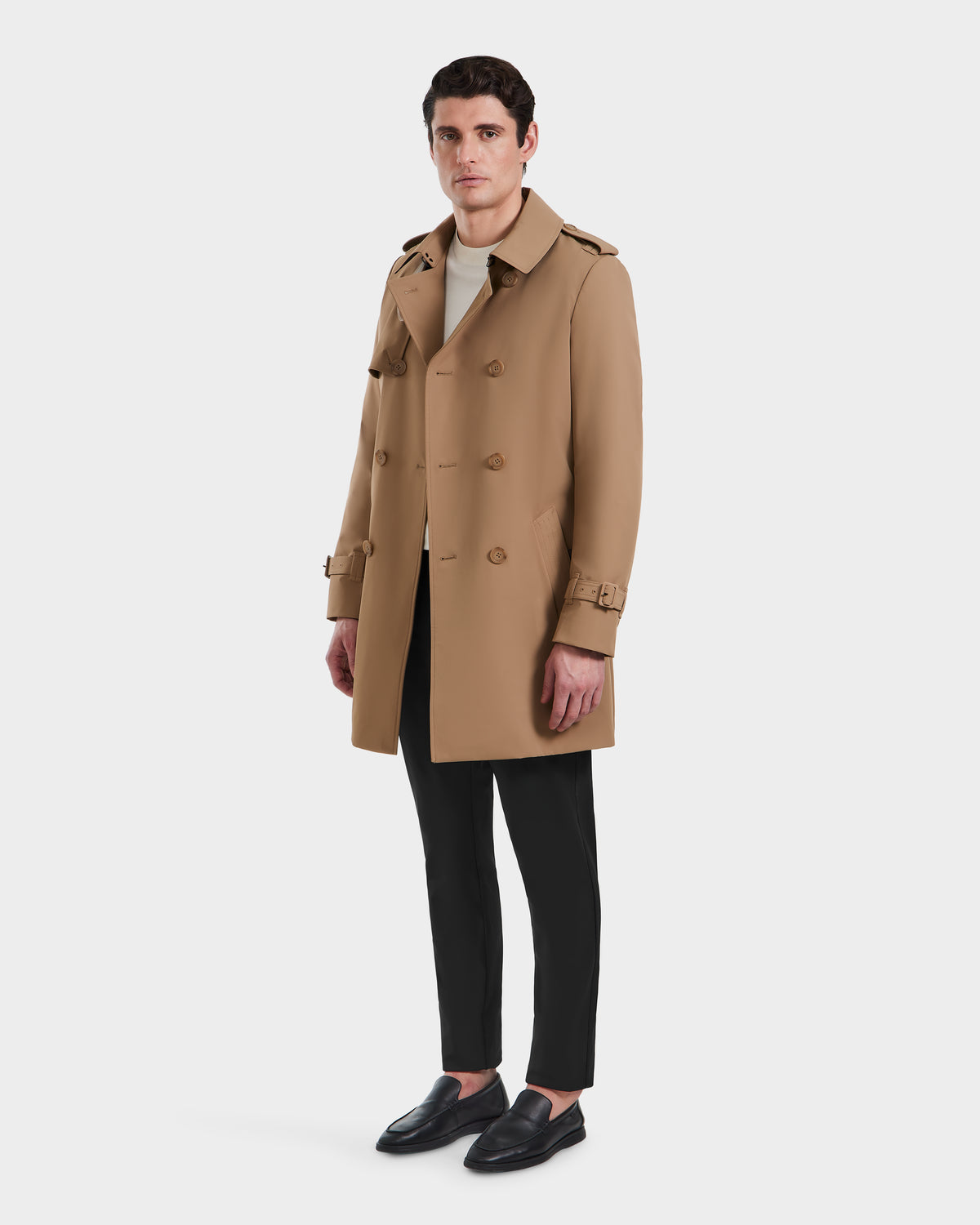 A man stands against a white background, wearing the Rudsak TIM - 6124025 Camel trench coat, black pants, a light-colored shirt, and black loafers.