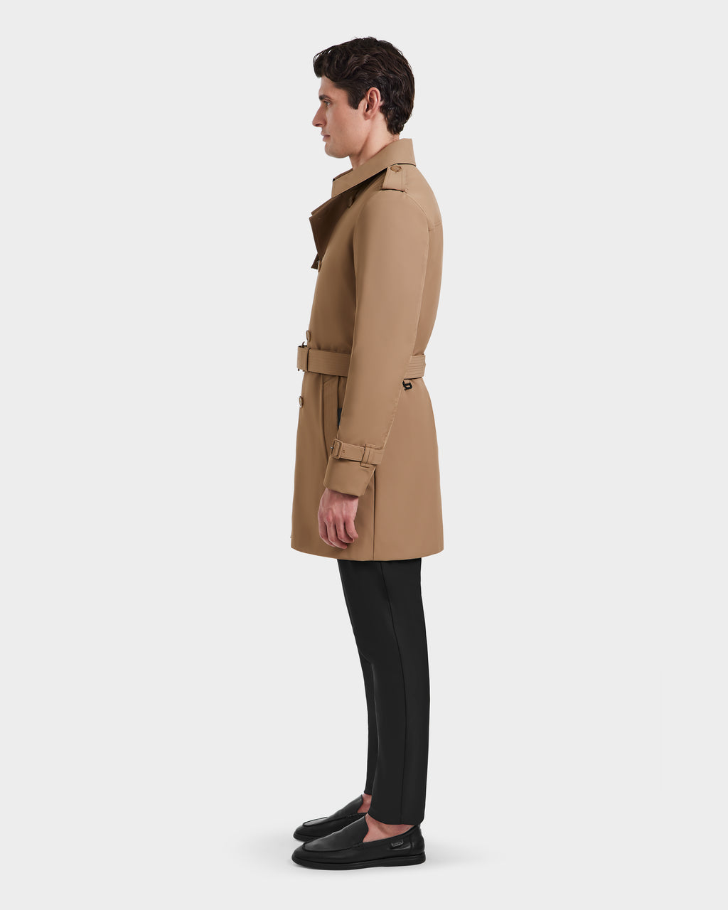 A man in profile facing left wears the Rudsak TIM - 6124025 Camel trench coat, belted, with black pants and loafers, standing against a plain light background.