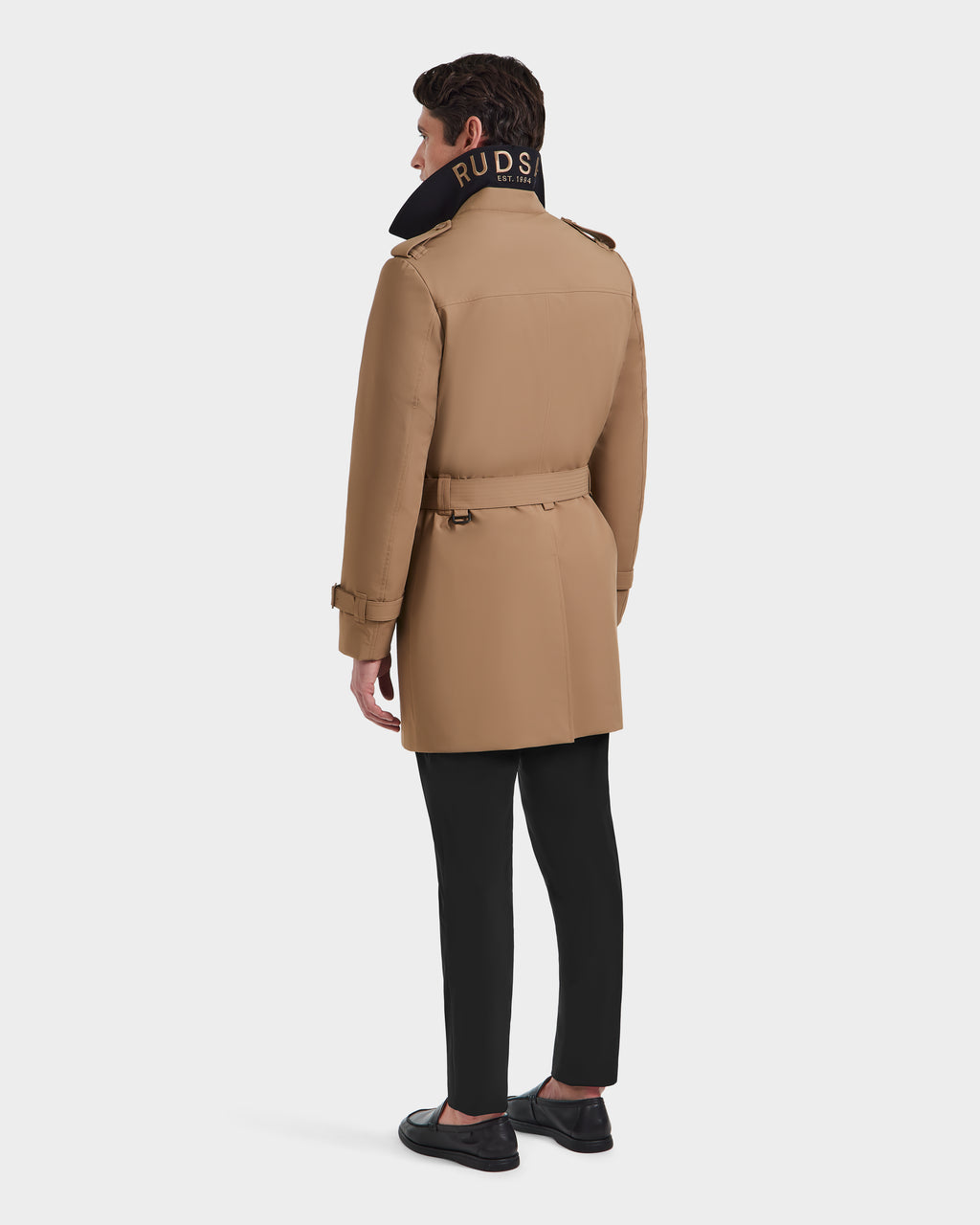 A man faces away wearing the Rudsak TIM - 6124025 Camel mens trench coat, featuring RUDSAK text on its raised black collar, paired with black pants and shoes, set against a plain white background.