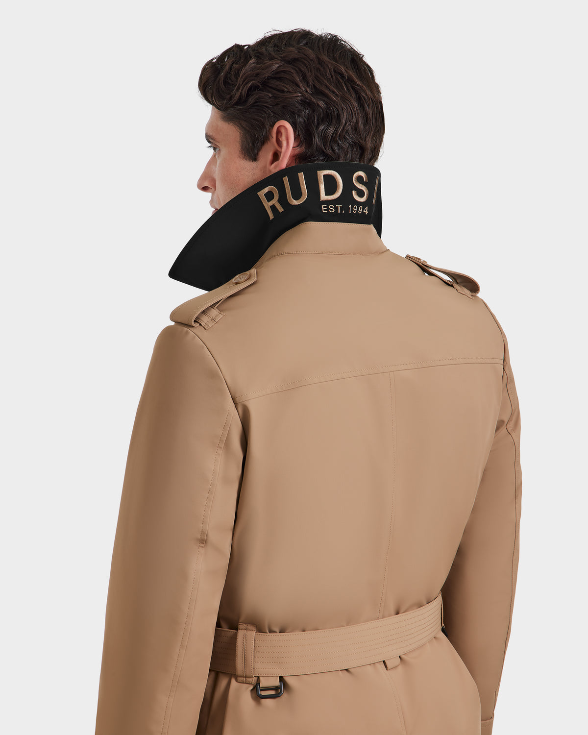 A man is shown from the back wearing the Rudsak TIM - 6124025 Camel trench coat. The collar is turned up, exposing black lining with gold text reading RUDSAK EST. 1994. The background is plain white.