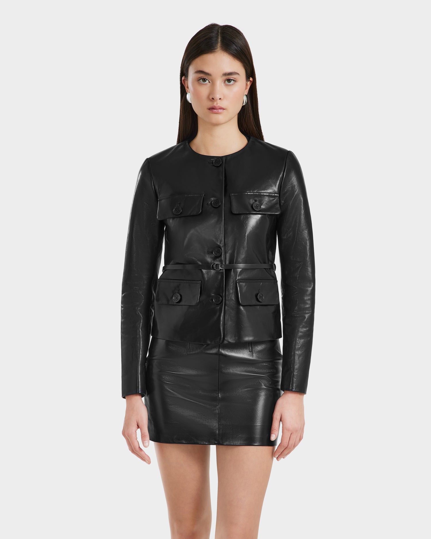 A woman with straight brown hair wears the Rudsak TRINITY - 8125022 Black cropped leather jacket and a matching black mini skirt, showcasing modern sophistication against a plain white background.