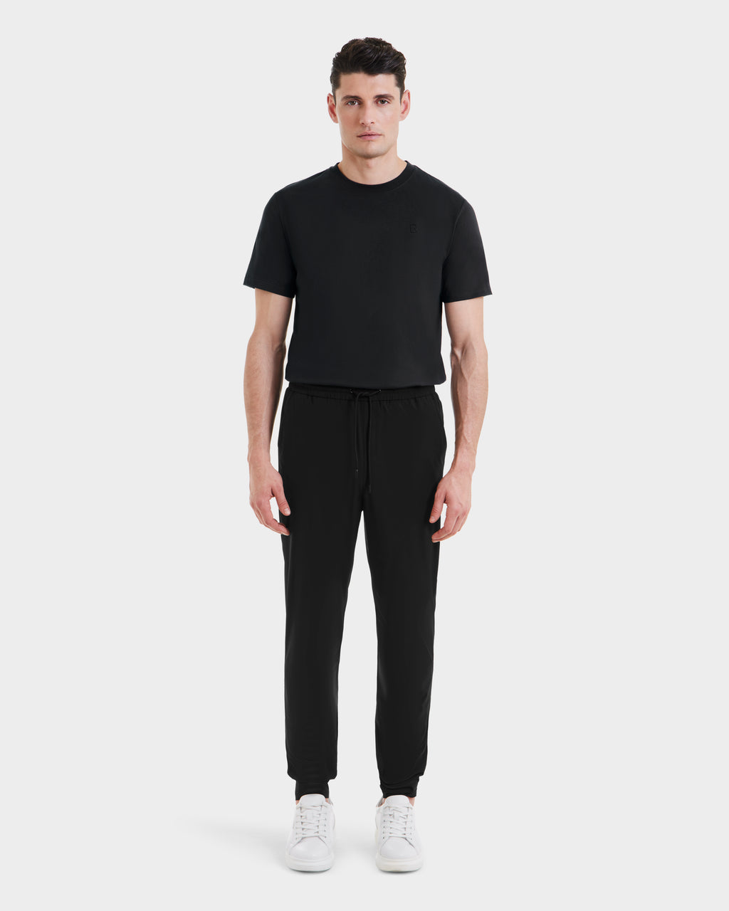 A man faces forward against a light gray background, wearing Rudsaks TUSTIN - 6525029 Black tailored-fit short-sleeve t-shirt and jogger pants with drawstring, paired with white sneakers—ideal for modern men’s casual style.