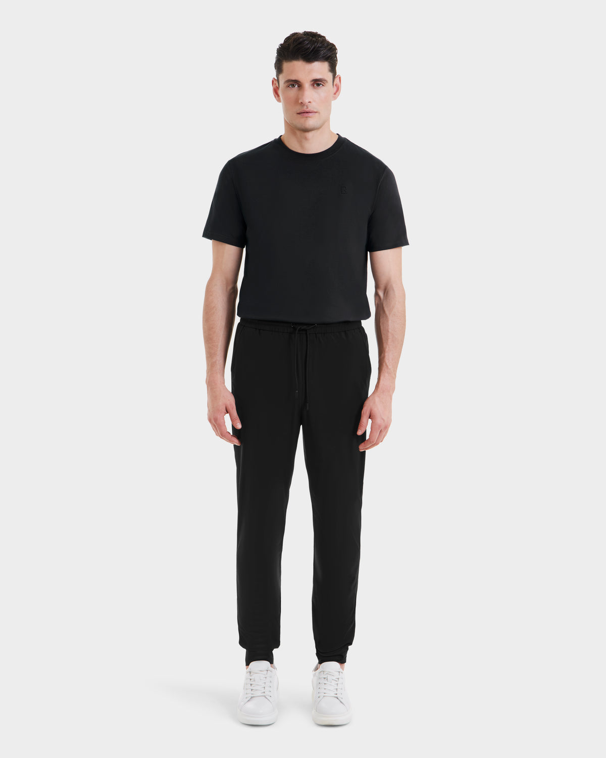 A man faces forward against a light gray background, wearing Rudsaks TUSTIN - 6525029 Black tailored-fit short-sleeve t-shirt and jogger pants with drawstring, paired with white sneakers—ideal for modern men’s casual style.