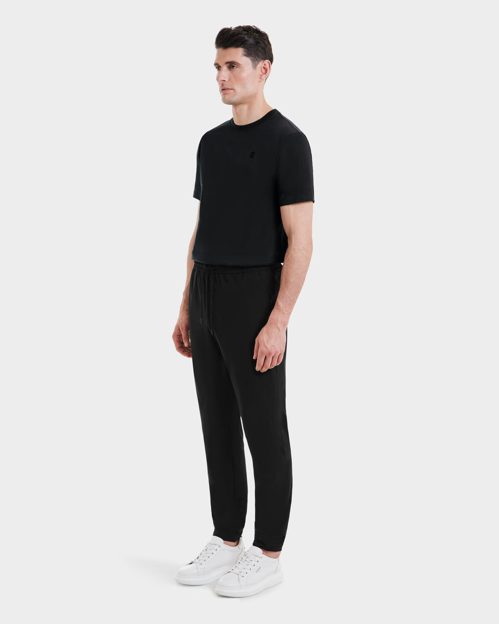 A man stands against a plain light background wearing Rudsaks TUSTIN - 6525029 Black tailored fit t-shirt and black jogger pants, paired with white sneakers. He faces slightly left, arms relaxed at his sides.