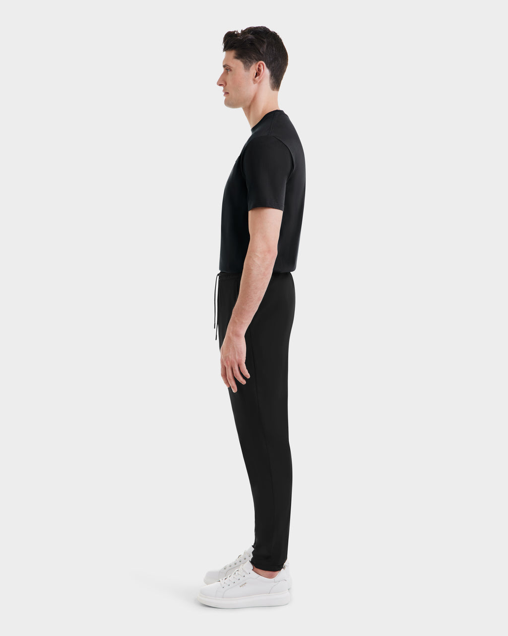 A man stands in profile facing left, wearing the Rudsak TUSTIN - 6525029 Black tailored fit T-shirt and matching black jogger pants with white sneakers—a sleek take on casual menswear against a plain white background.