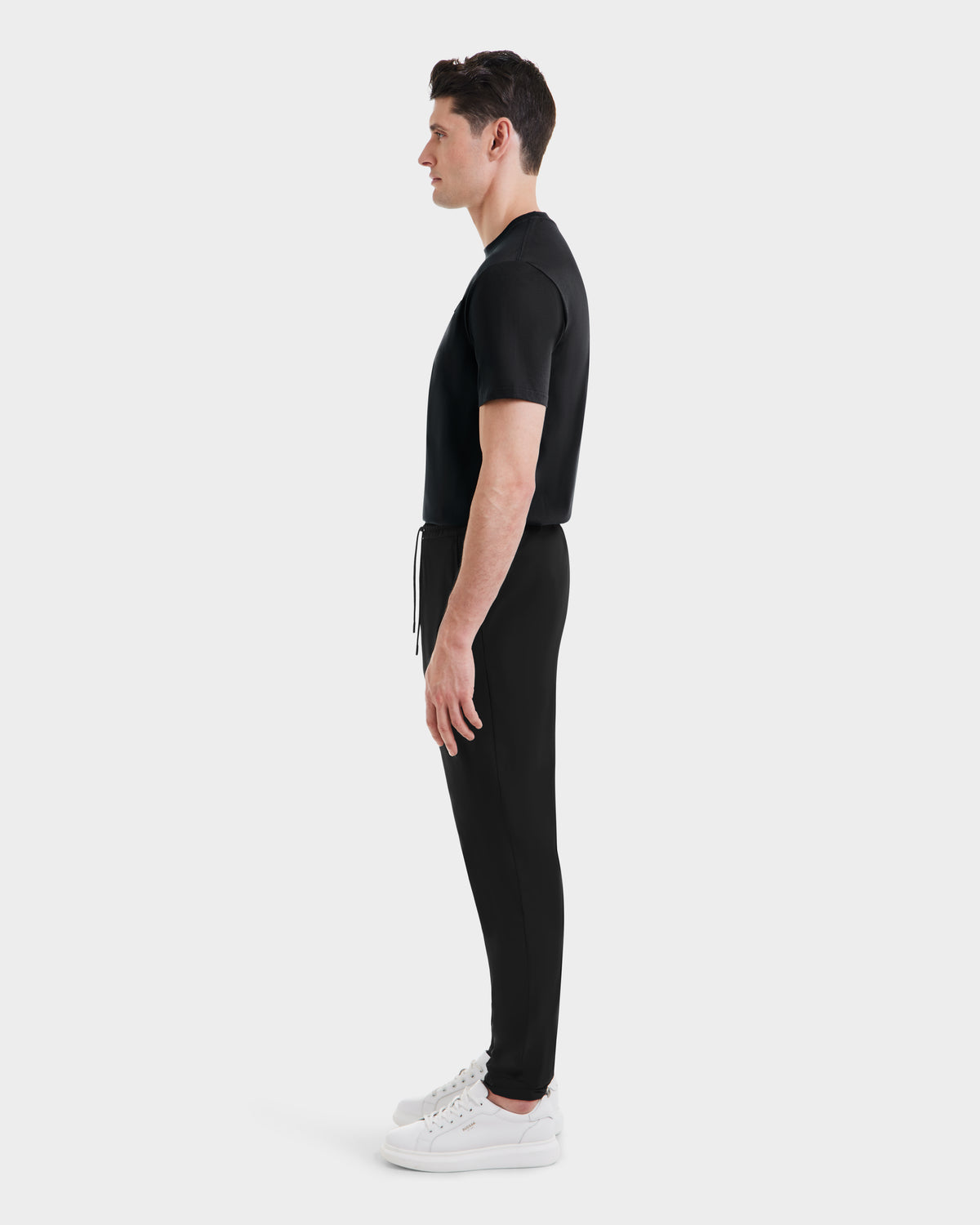 A man stands in profile facing left, wearing the Rudsak TUSTIN - 6525029 Black tailored fit T-shirt and matching black jogger pants with white sneakers—a sleek take on casual menswear against a plain white background.