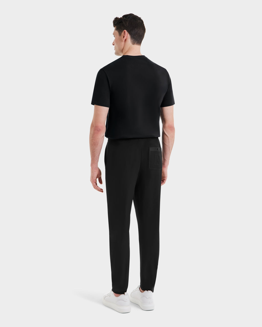 A man stands facing away, wearing Rudsak’s TUSTIN - 6525029 Black short-sleeve shirt and tailored jogger pants with white sneakers against a plain light gray background—an effortless mens casual look.