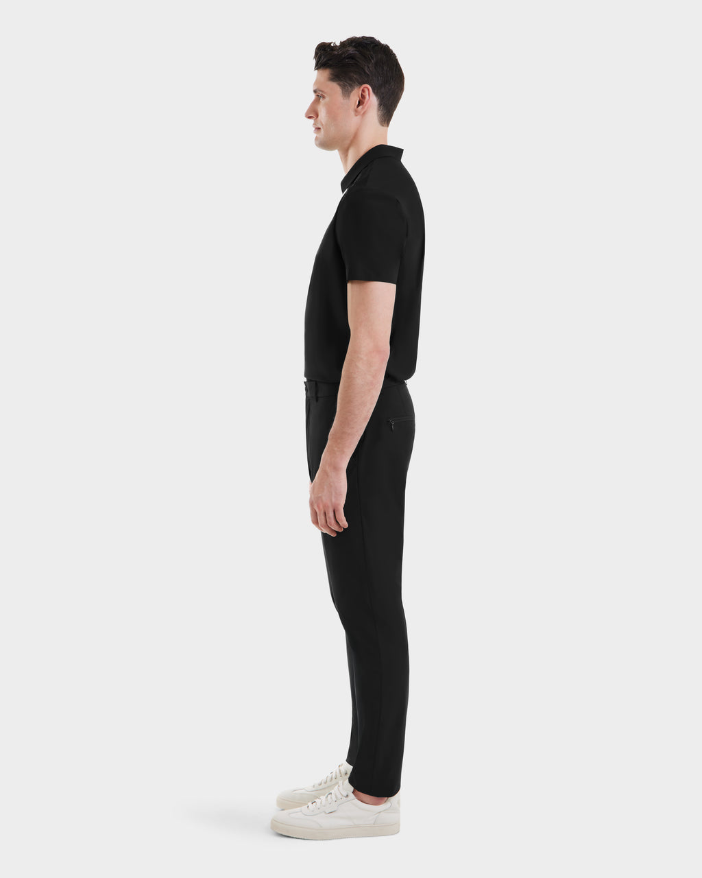 A man in profile faces left, wearing Rudsak’s VERNON - 6525025 Black short-sleeve polo and tapered technical trousers with white sneakers, set against a plain white backdrop—ideal for the modern wardrobe.