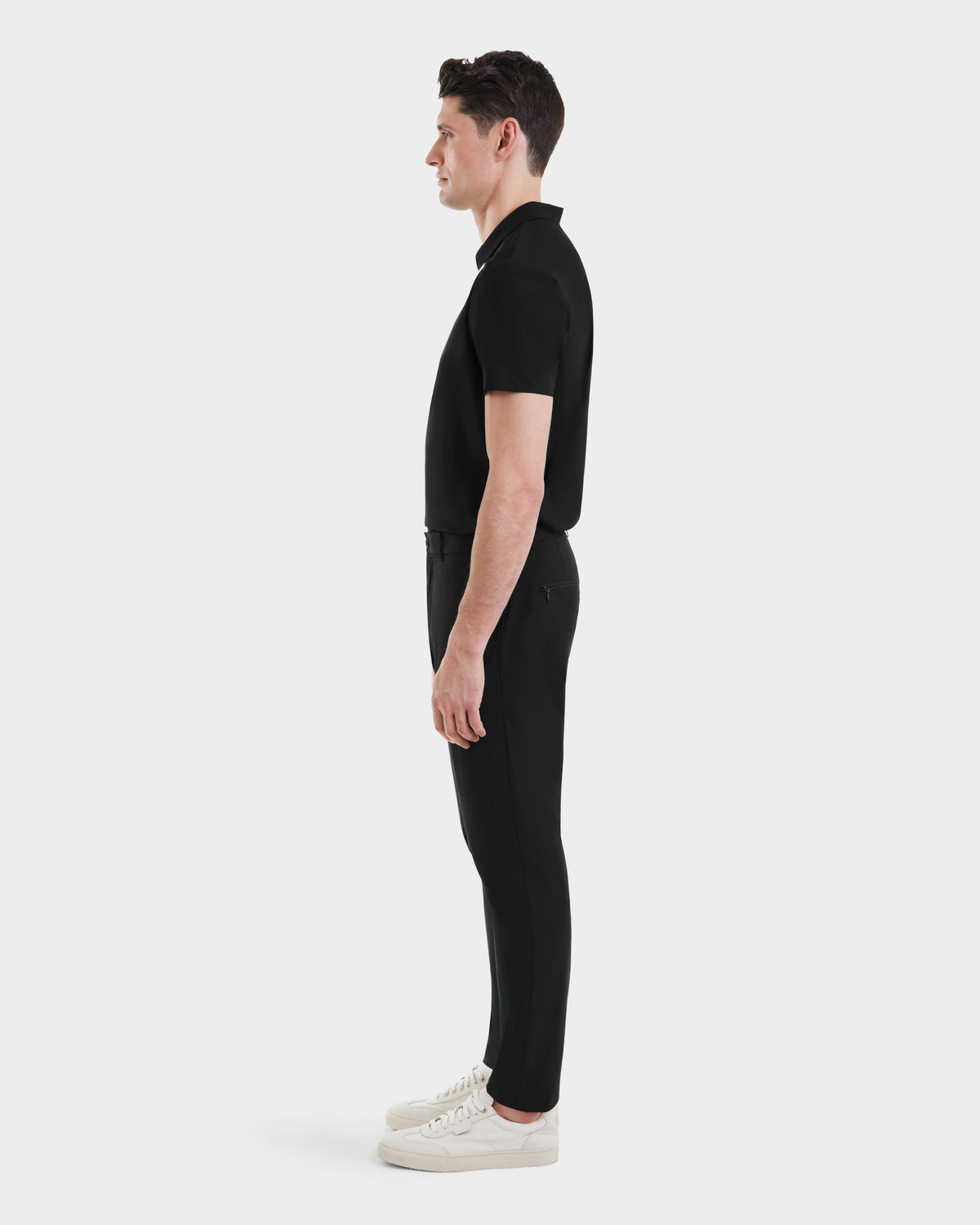 A man in profile faces left, wearing Rudsak’s VERNON - 6525025 Black short-sleeve polo and tapered technical trousers with white sneakers, set against a plain white backdrop—ideal for the modern wardrobe.