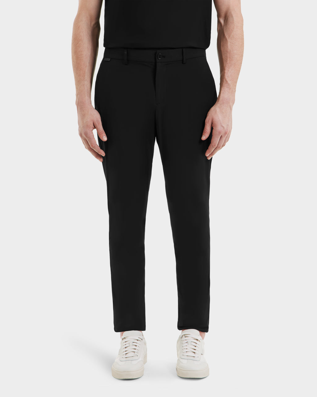 A person stands facing forward against a plain white background, shown from the shoulders down, wearing Rudsak VERNON - 6525025 Black tapered-leg trousers and a black shirt paired with white sneakers.
