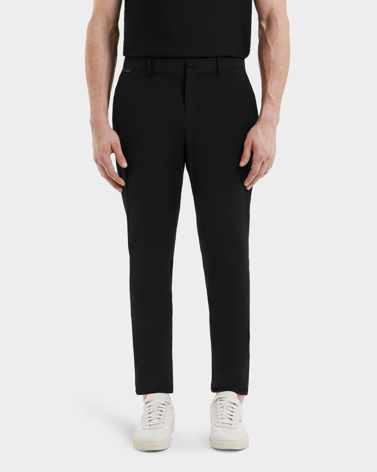A person stands facing forward against a plain white background, shown from the shoulders down, wearing Rudsak VERNON - 6525025 Black tapered-leg trousers and a black shirt paired with white sneakers.
