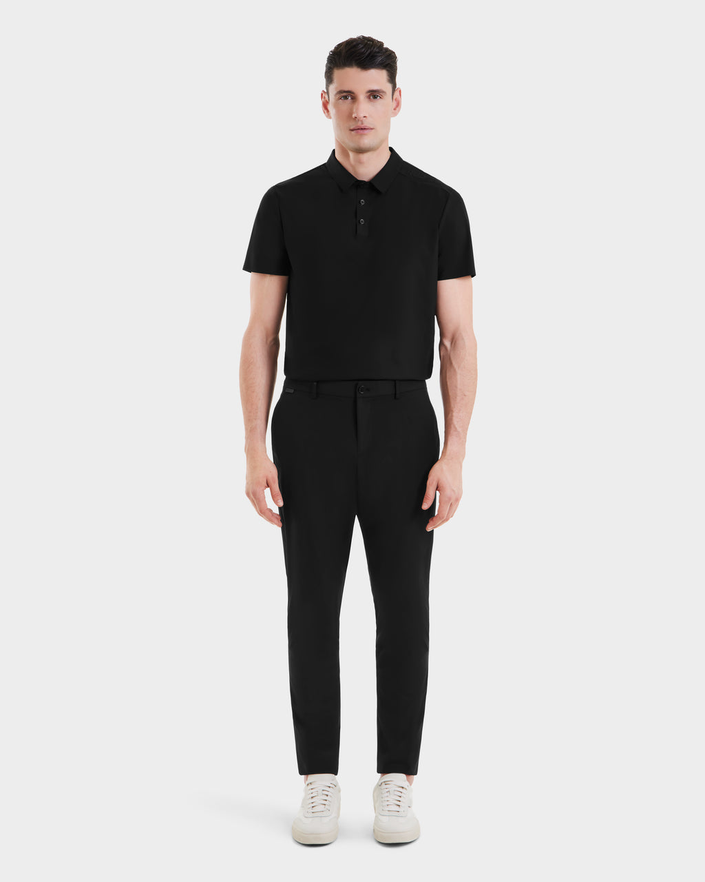 A man faces forward against a white background wearing the VERNON - 6525025 Black polo shirt by Rudsak, styled with tapered trousers and white sneakers for an effortlessly modern look.