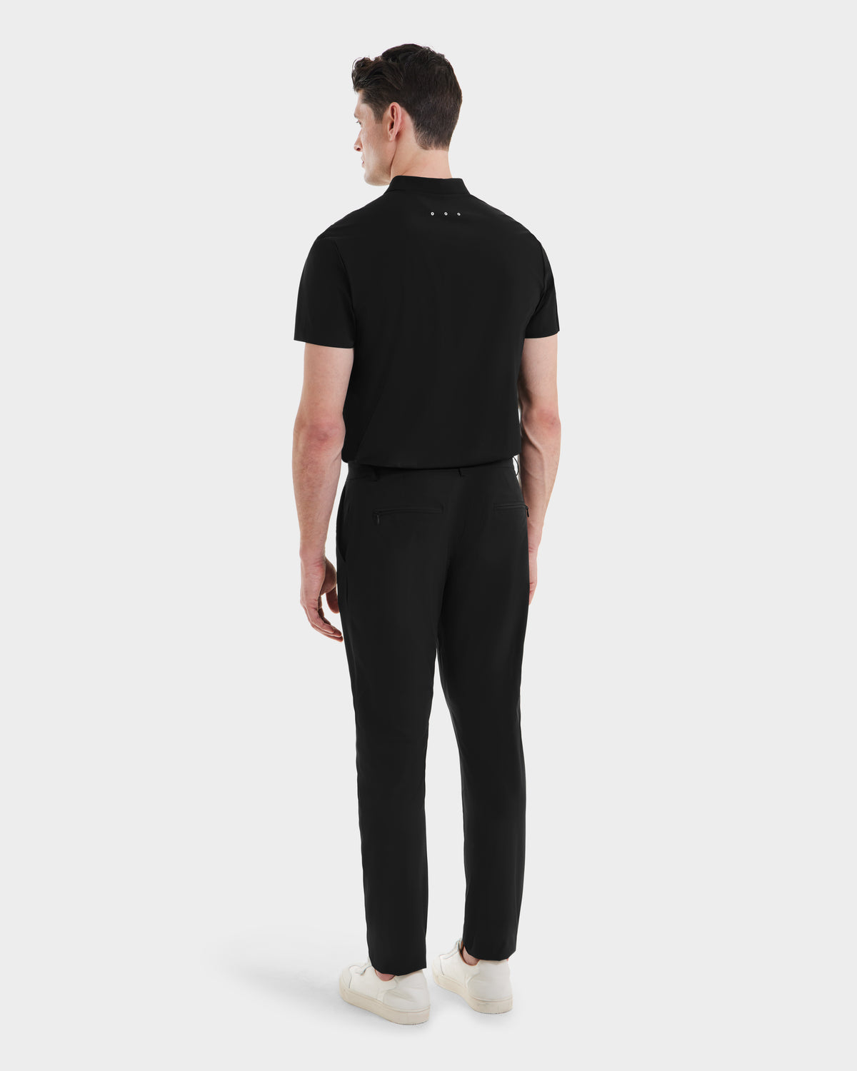 A man stands facing away in Rudsaks VERNON - 6525025 Black polo shirt, paired with black tapered-leg trousers and white sneakers against a white background—a sleek, modern wardrobe choice.
