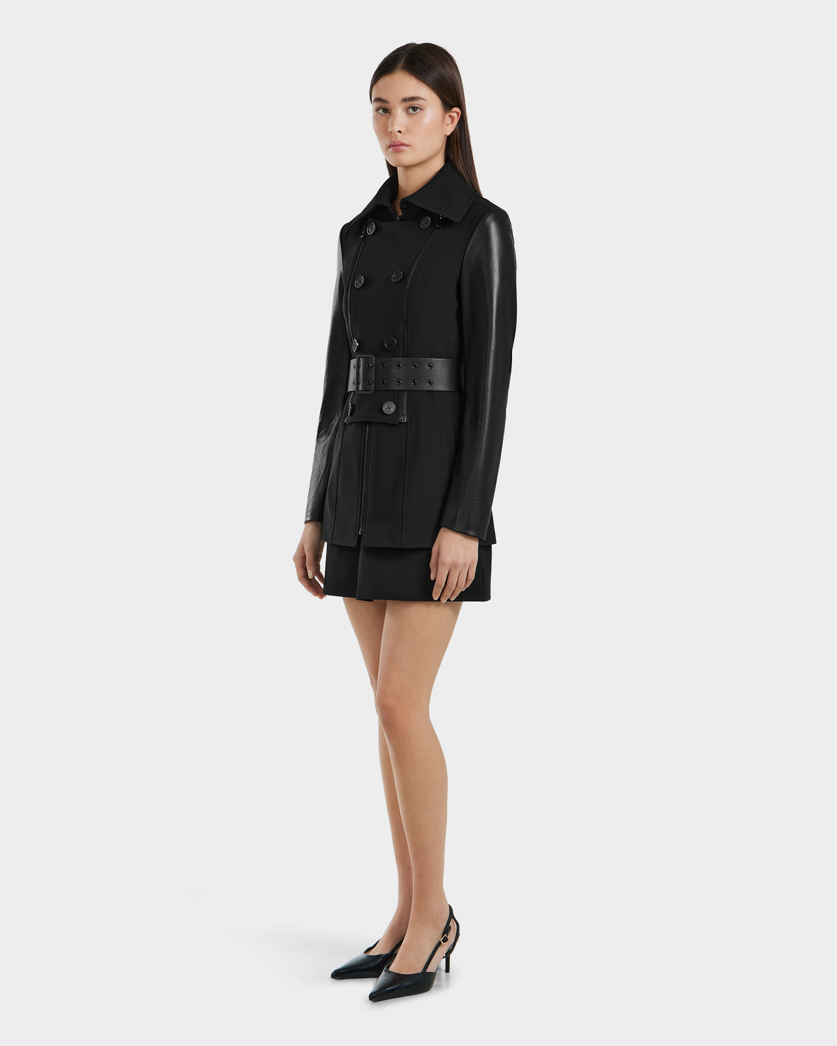A woman stands against a plain background wearing the Rudsak VIRGINIE S - 8125053 Black trench coat and black heels, her hair down, hands by her sides, looking forward with a neutral expression.