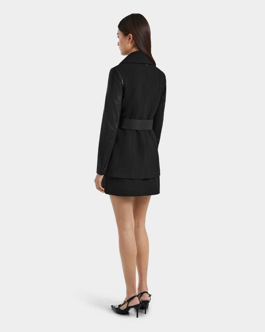 A woman with straight brown hair faces away, wearing the VIRGINIE S - 8125053 Black tailored trench coat by Rudsak, styled as a belted blazer with a short black skirt and heels against a plain light background.