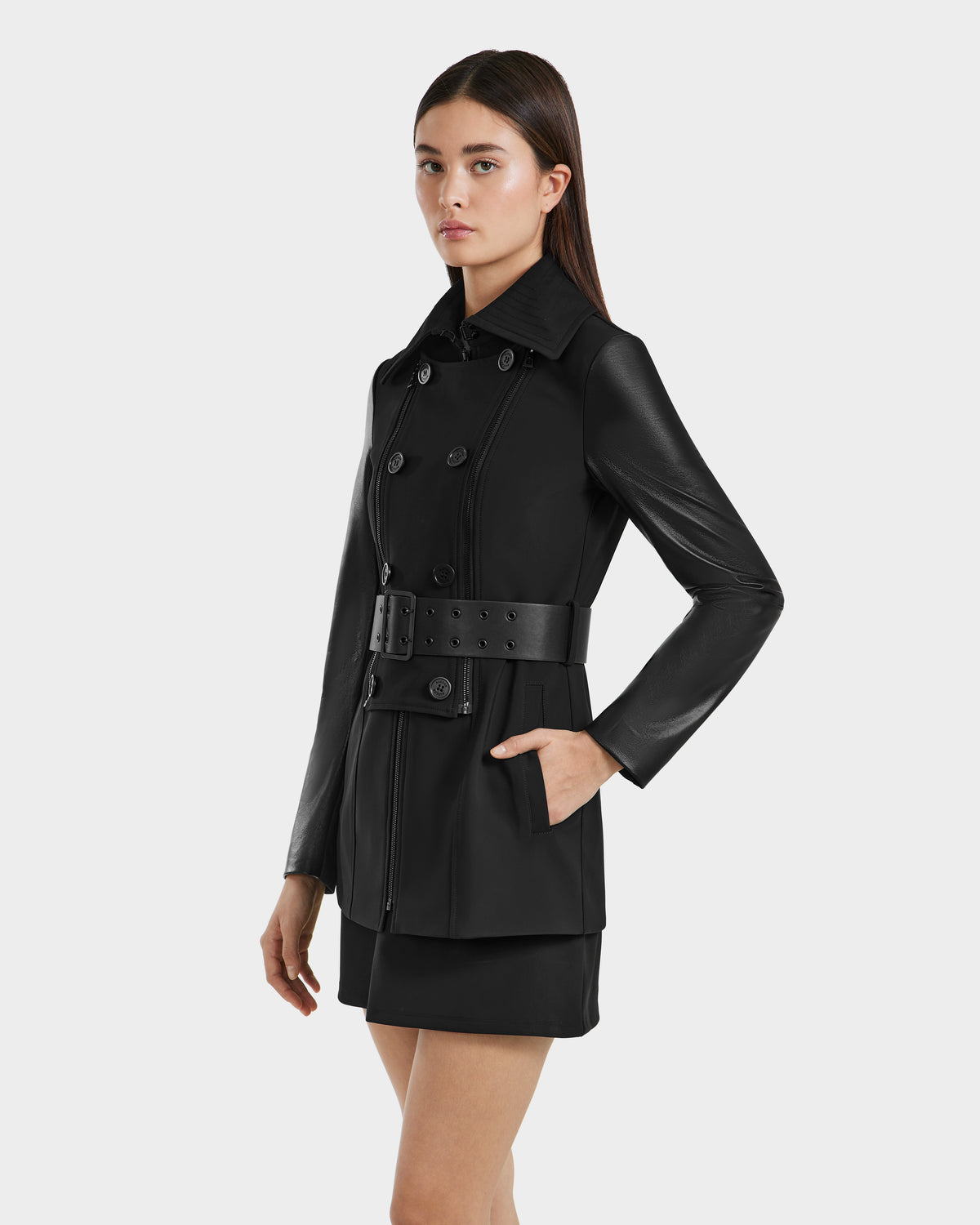 A woman with straight dark hair and a neutral expression wears the Rudsak VIRGINIE S - 8125053 Black tailored trench coat and black skirt, standing left against a plain light gray background.