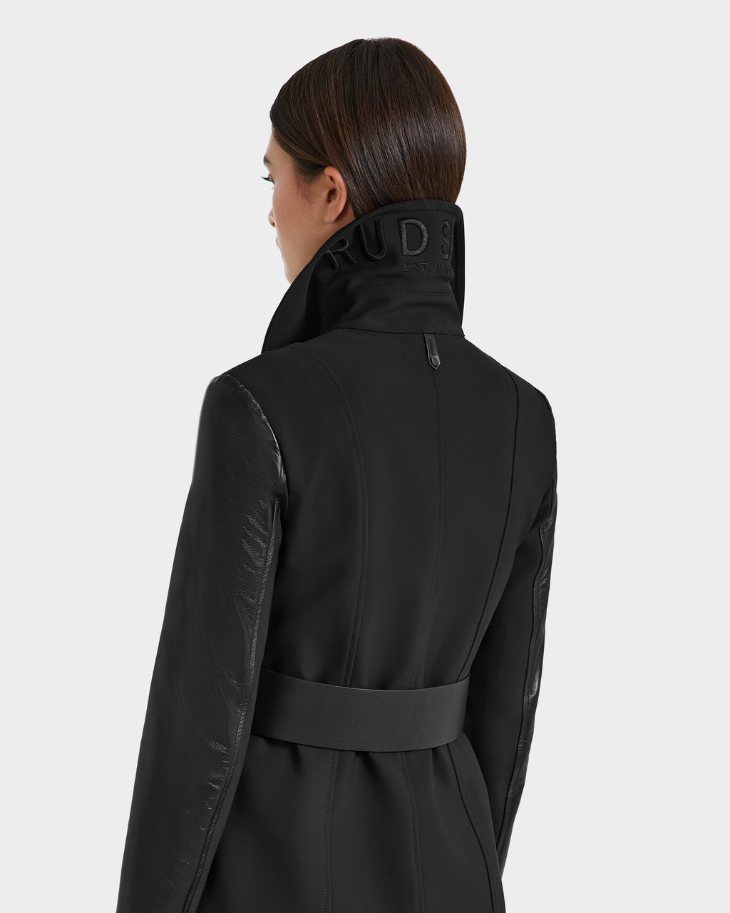 A woman with straight brown hair wears the VIRGINIE S - 8125053 Black trench coat by Rudsak, featuring a high belted collar with the partially visible RUDS logo, viewed from the back against a plain light gray background.