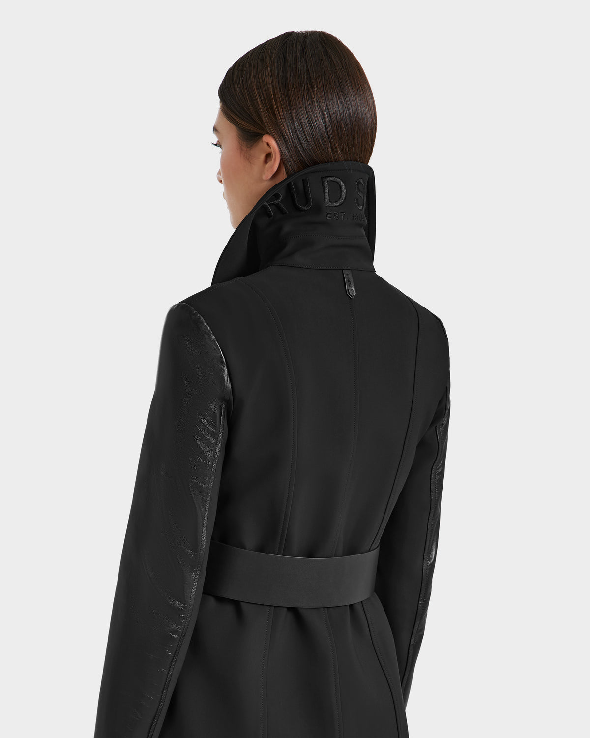 A woman with straight brown hair wears the VIRGINIE S - 8125053 Black trench coat by Rudsak, featuring a high belted collar with the partially visible RUDS logo, viewed from the back against a plain light gray background.