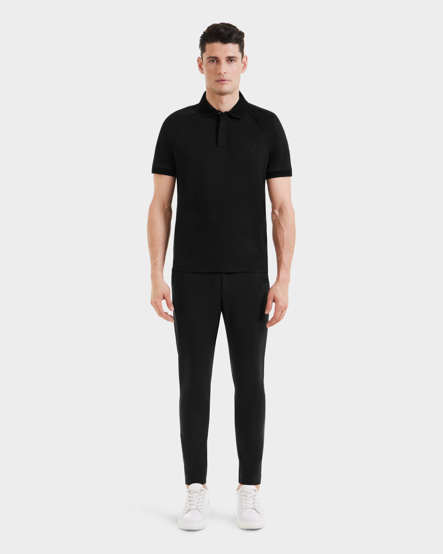 A man stands facing forward wearing the WALTER - 6524531 Black/Black polo by Rudsak, paired with black pants and white sneakers against a plain light background.