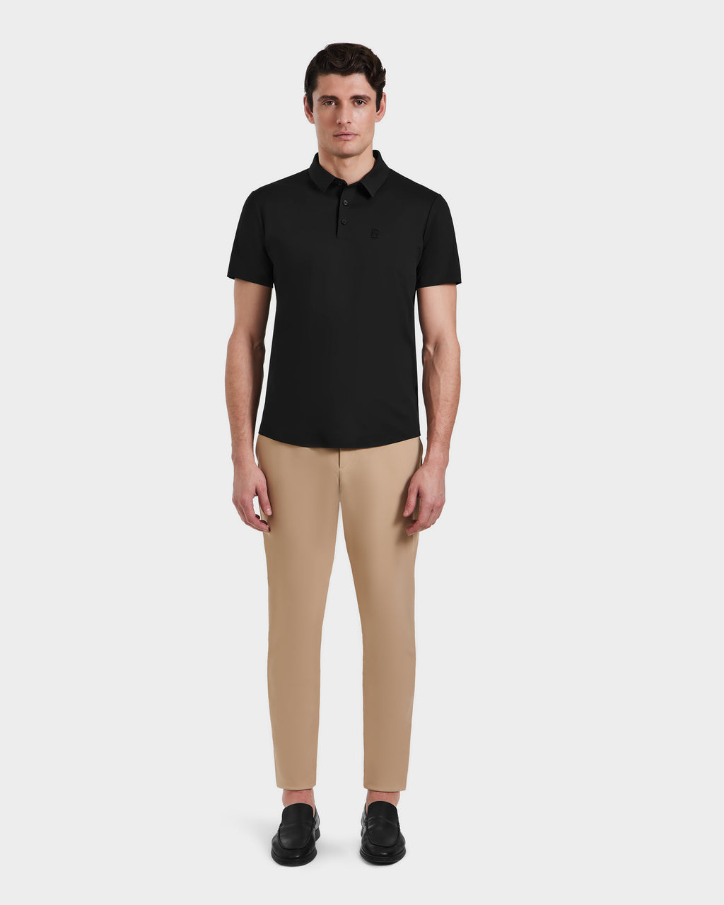 A man stands facing forward against a plain white background, wearing Rudsaks WELLS - 6525017 Black short-sleeve polo shirt, styled with tan slim-fit technical pants and black shoes—modern essentials for men.