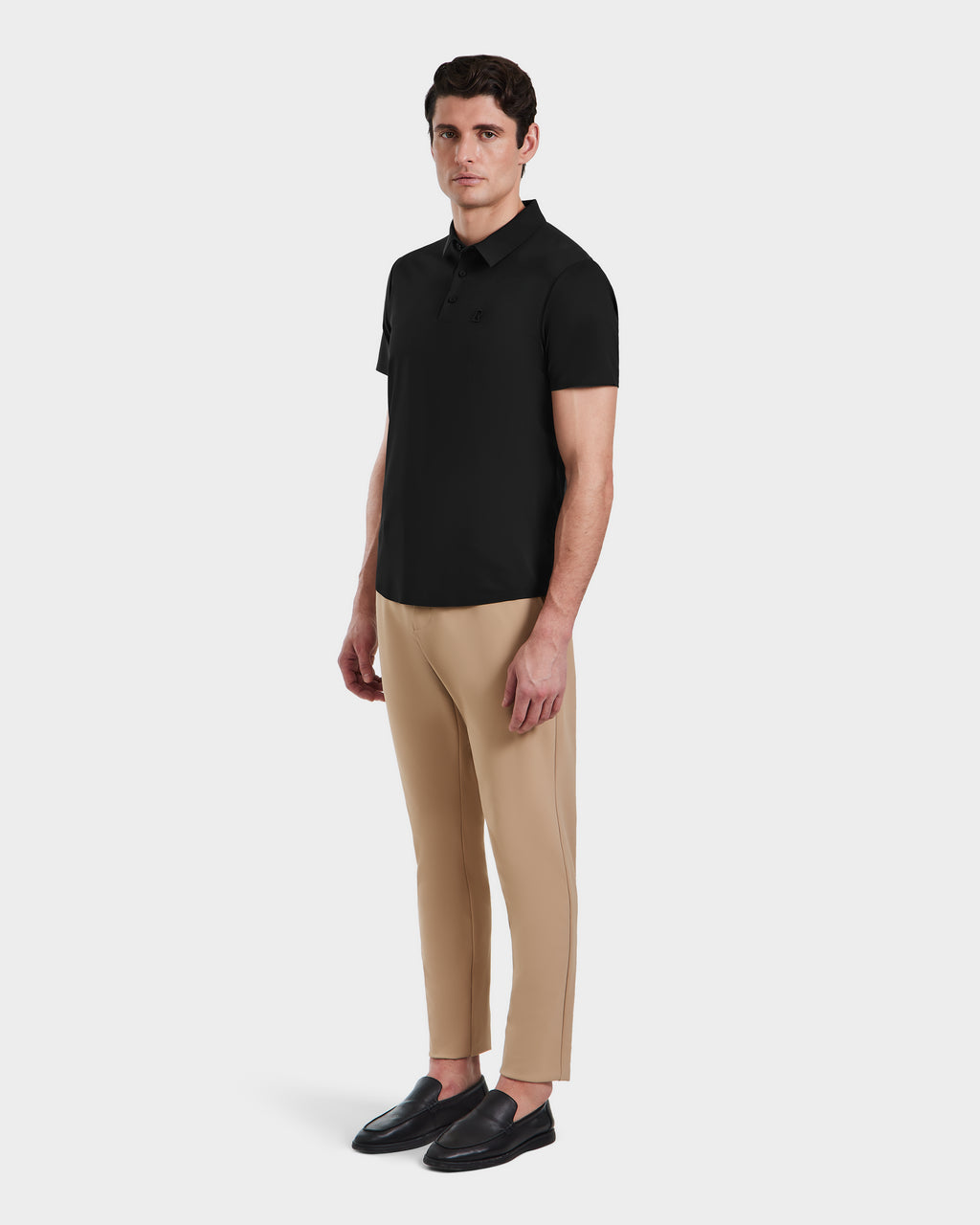 A man models the Rudsak WELLS - 6525017 Black short-sleeve polo, featuring lightweight technical fabric. He pairs it with beige pants and black loafers, standing against a plain background with a neutral expression.