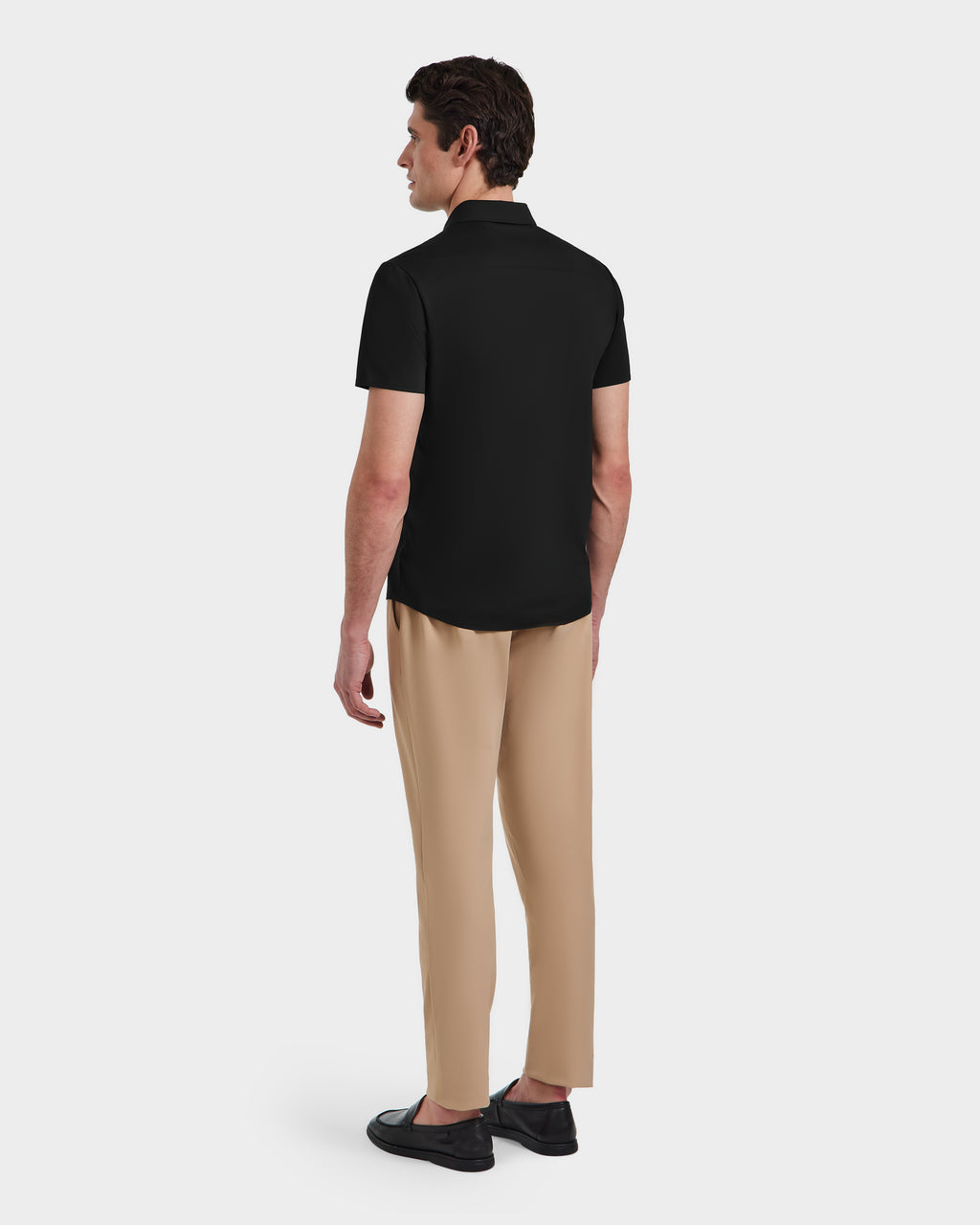 A man faces away, wearing the Rudsak WELLS - 6525017 Black tailored-fit short-sleeve polo shirt with beige trousers and black shoes, set against a plain white background.