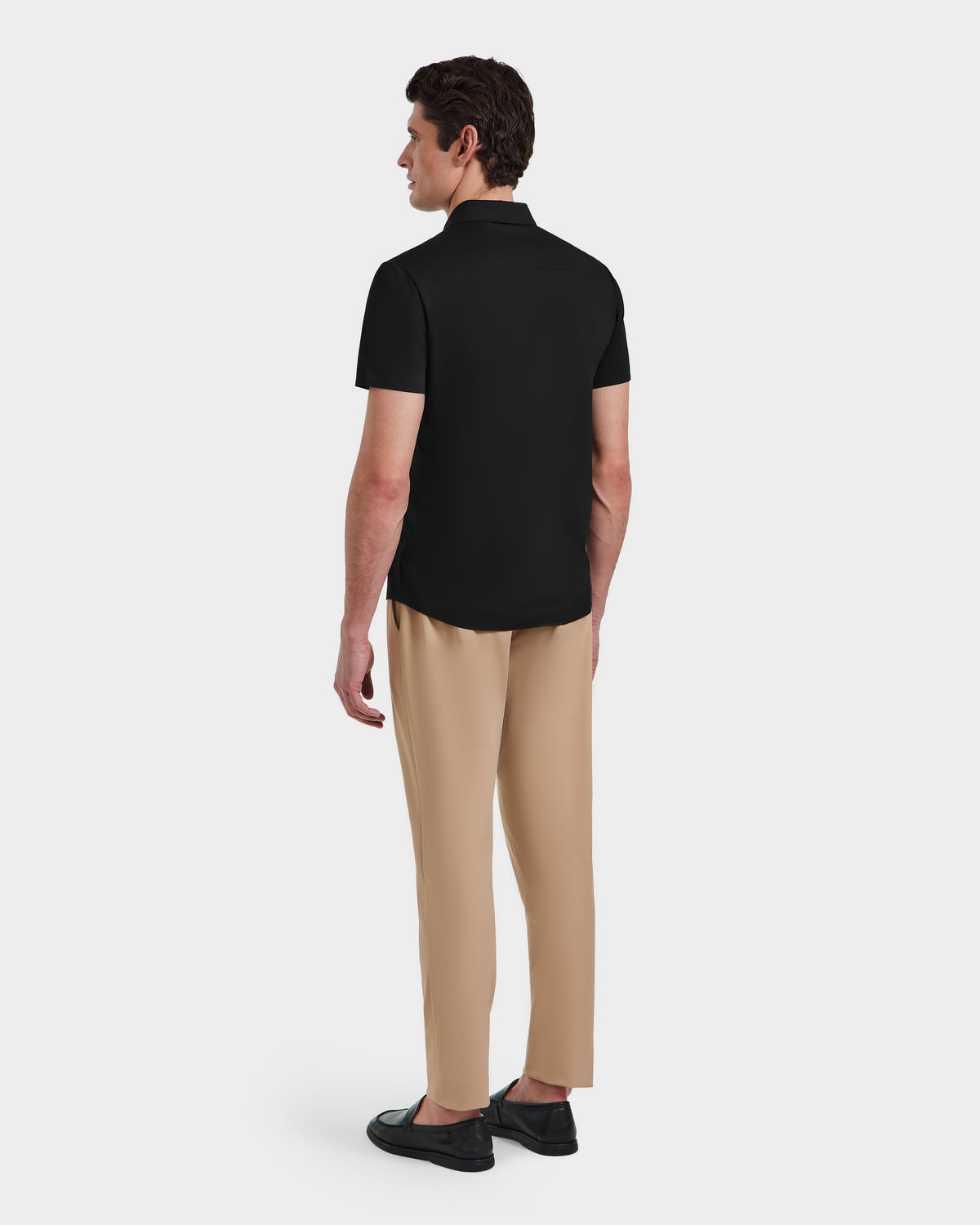 A man faces away, wearing the Rudsak WELLS - 6525017 Black tailored-fit short-sleeve polo shirt with beige trousers and black shoes, set against a plain white background.