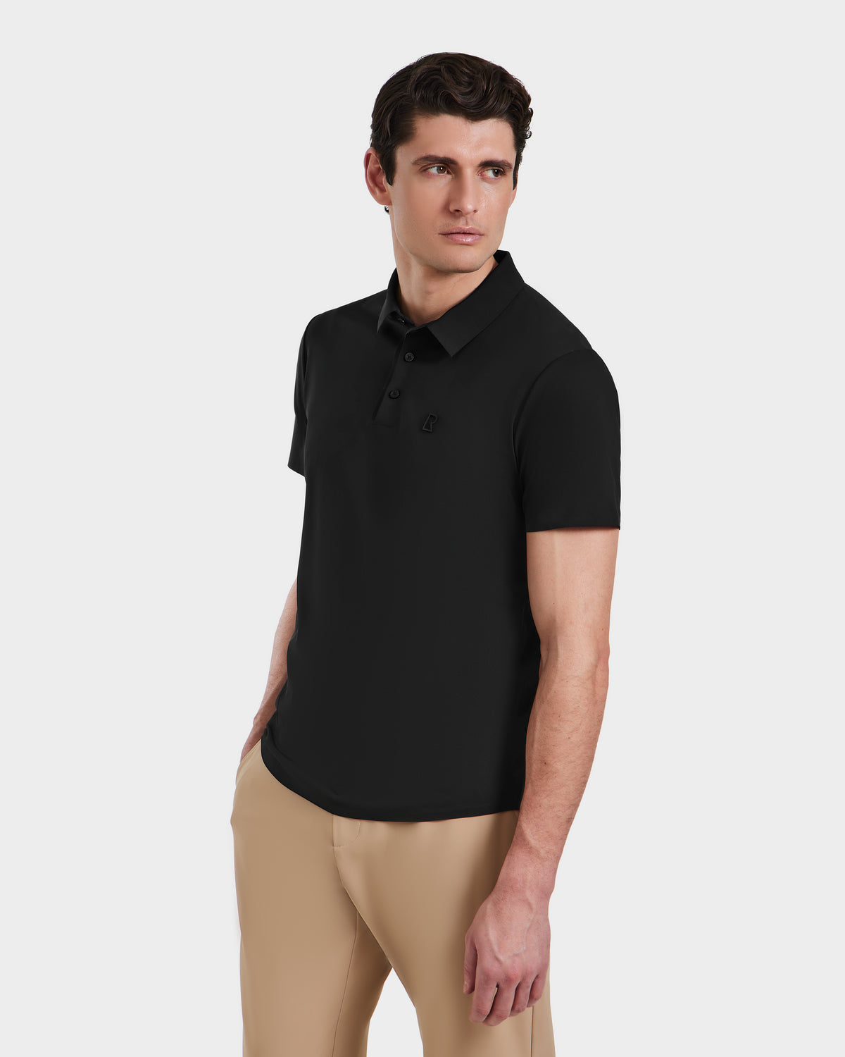 A man stands against a plain white background, wearing the Rudsak WELLS - 6525017 Black short-sleeve polo shirt in lightweight technical fabric and beige pants, with one hand in his pocket and looking to the side.