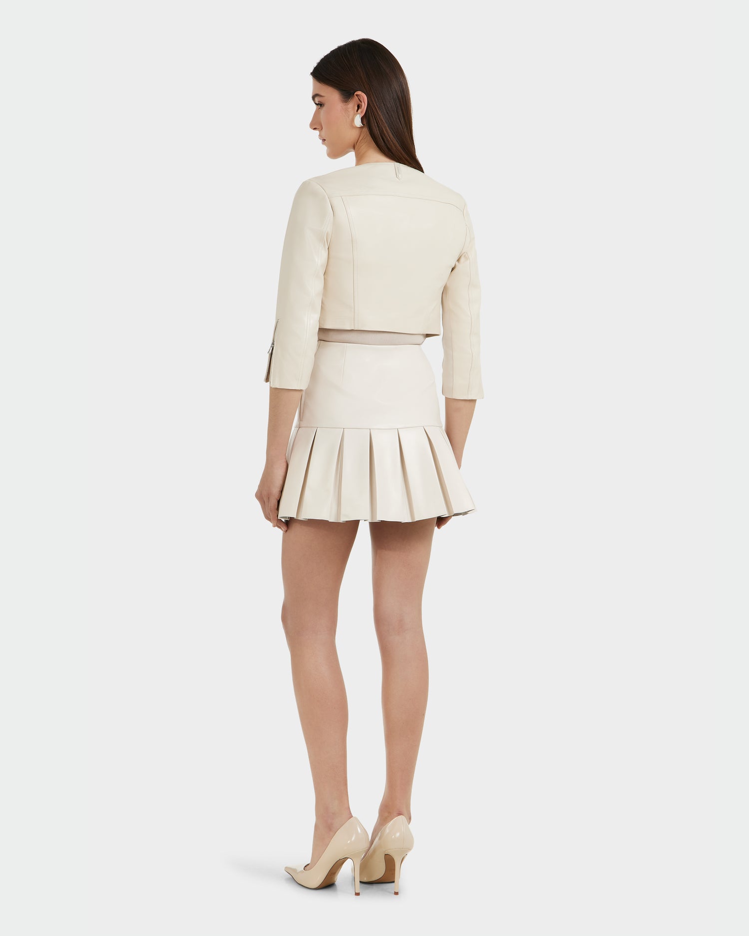 A woman stands with her back to the camera in the WINNI - 8124053 Vanilla cropped leather jacket by Rudsak, paired with a pleated mini skirt and nude high heels, against a plain white background.
