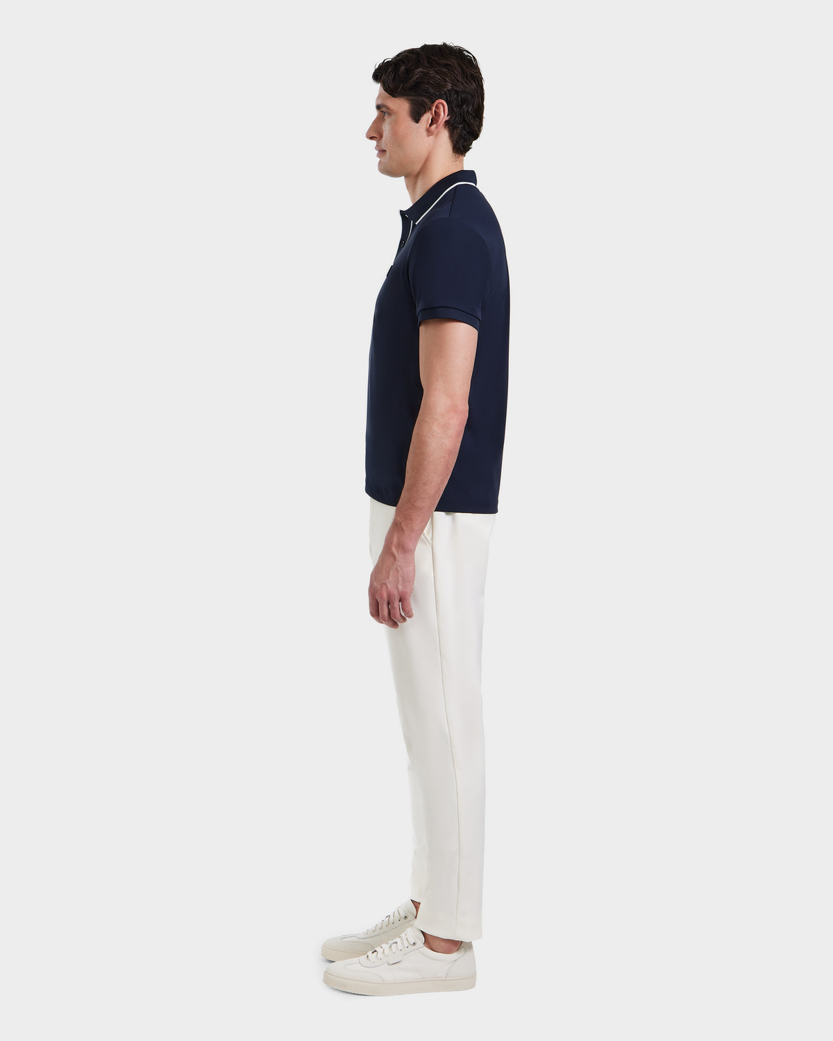 A man stands in profile against a plain background, wearing the Rudsak XAVIER - 6524027 Navy mens polo shirt, paired with white pants and white sneakers.