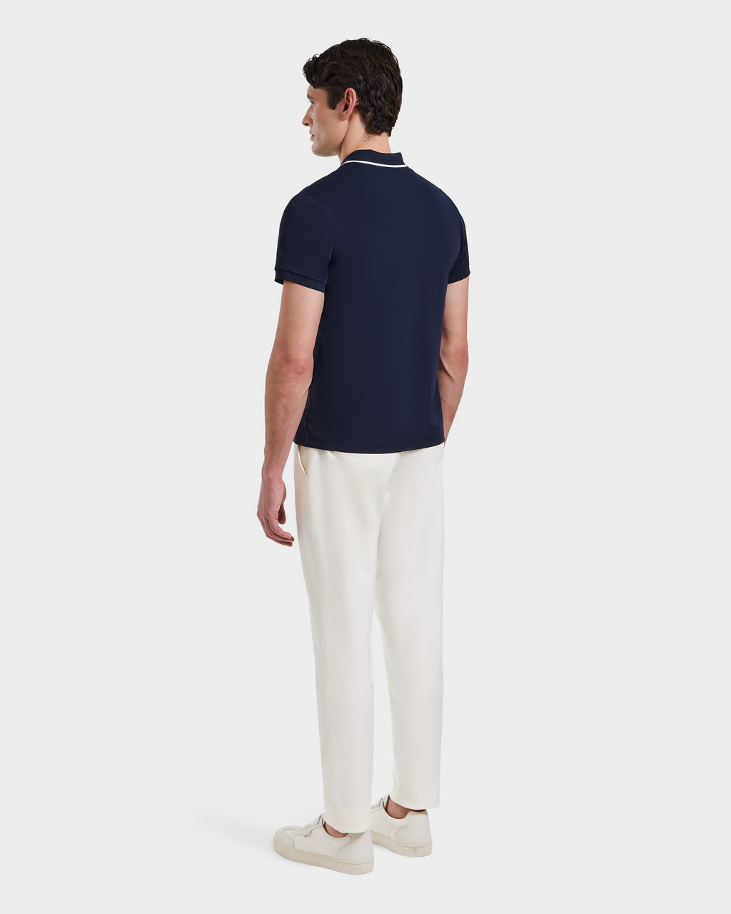 A man faces away, wearing the Rudsak XAVIER - 6524027 Navy polo shirt with white pants and sneakers, set against a plain white background.