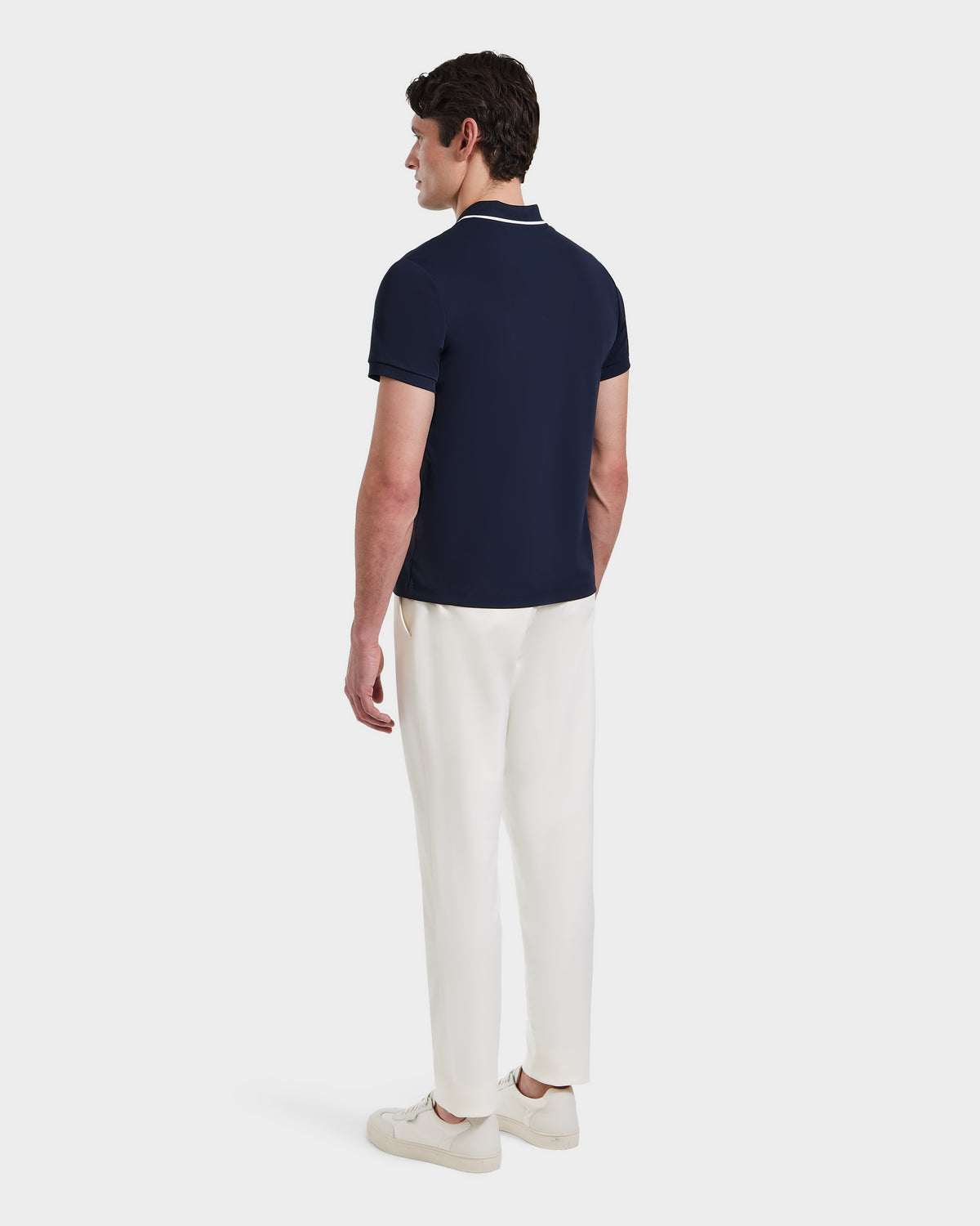 A man faces away, wearing the Rudsak XAVIER - 6524027 Navy polo shirt with white pants and sneakers, set against a plain white background.