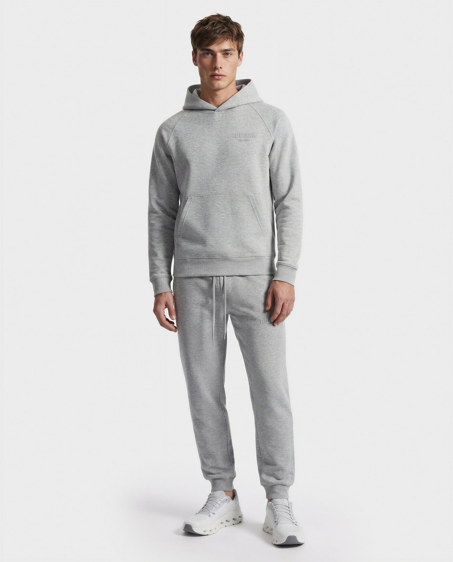 A young man faces forward, wearing the Rudsak BRIAR - 9525001 hoodie in Heather Grey (M), paired with matching jogger pants and white sneakers, set against a plain white background.