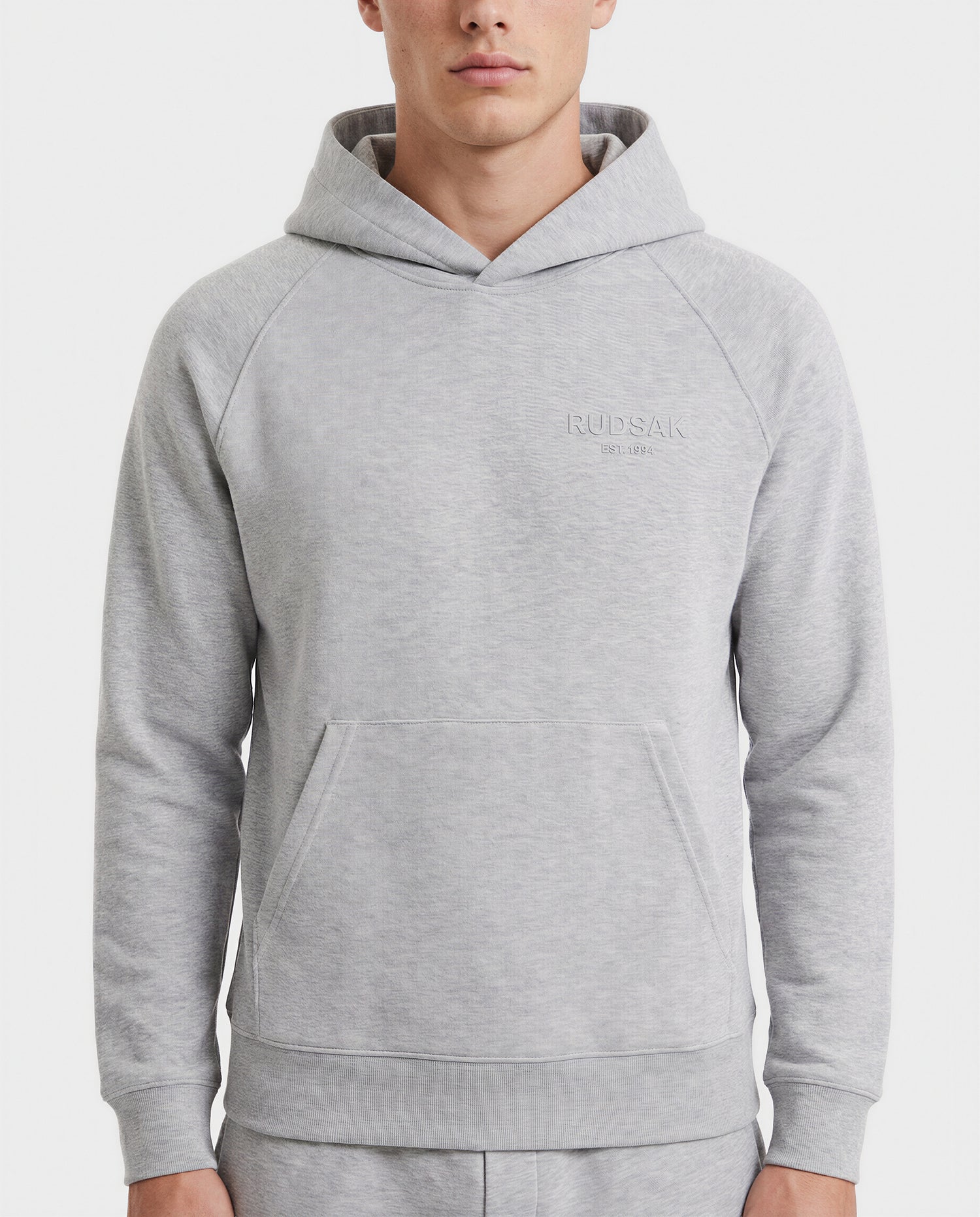 A person wears the Rudsak BRIAR - 9525001 hoodie in heather grey (M), a unisex premium cotton blend with a front pocket and Rudsak embroidered on the left chest, standing against a white background for a casual look.