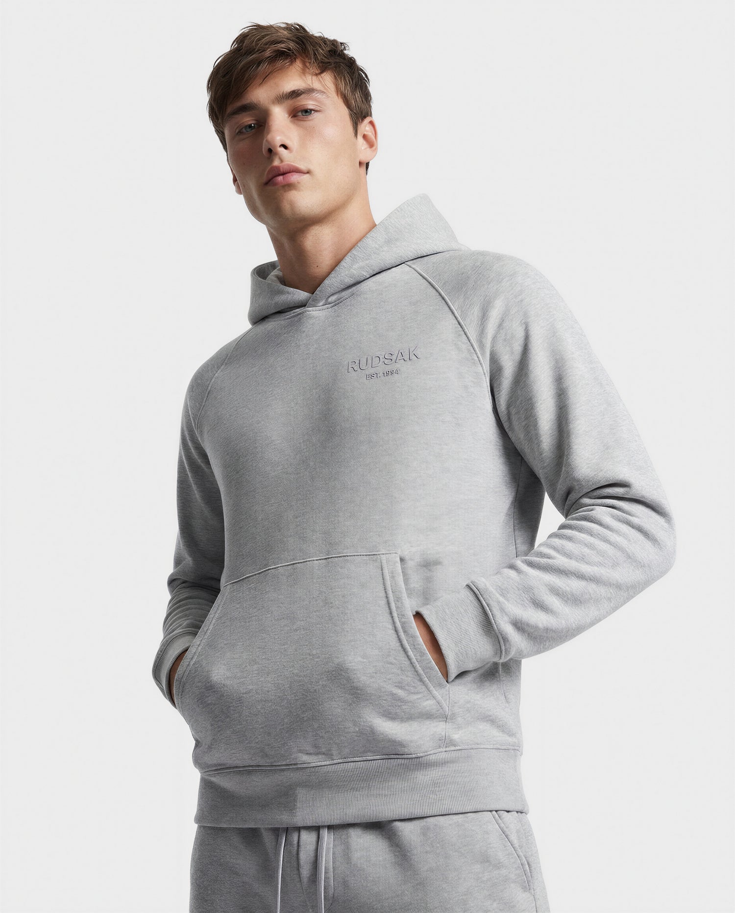 A young person with short brown hair poses neutrally against a white background, wearing the Rudsak BRIAR - 9525001 unisex hoodie in Heather Grey M, made from a premium cotton blend with a front pocket and subtle chest branding.
