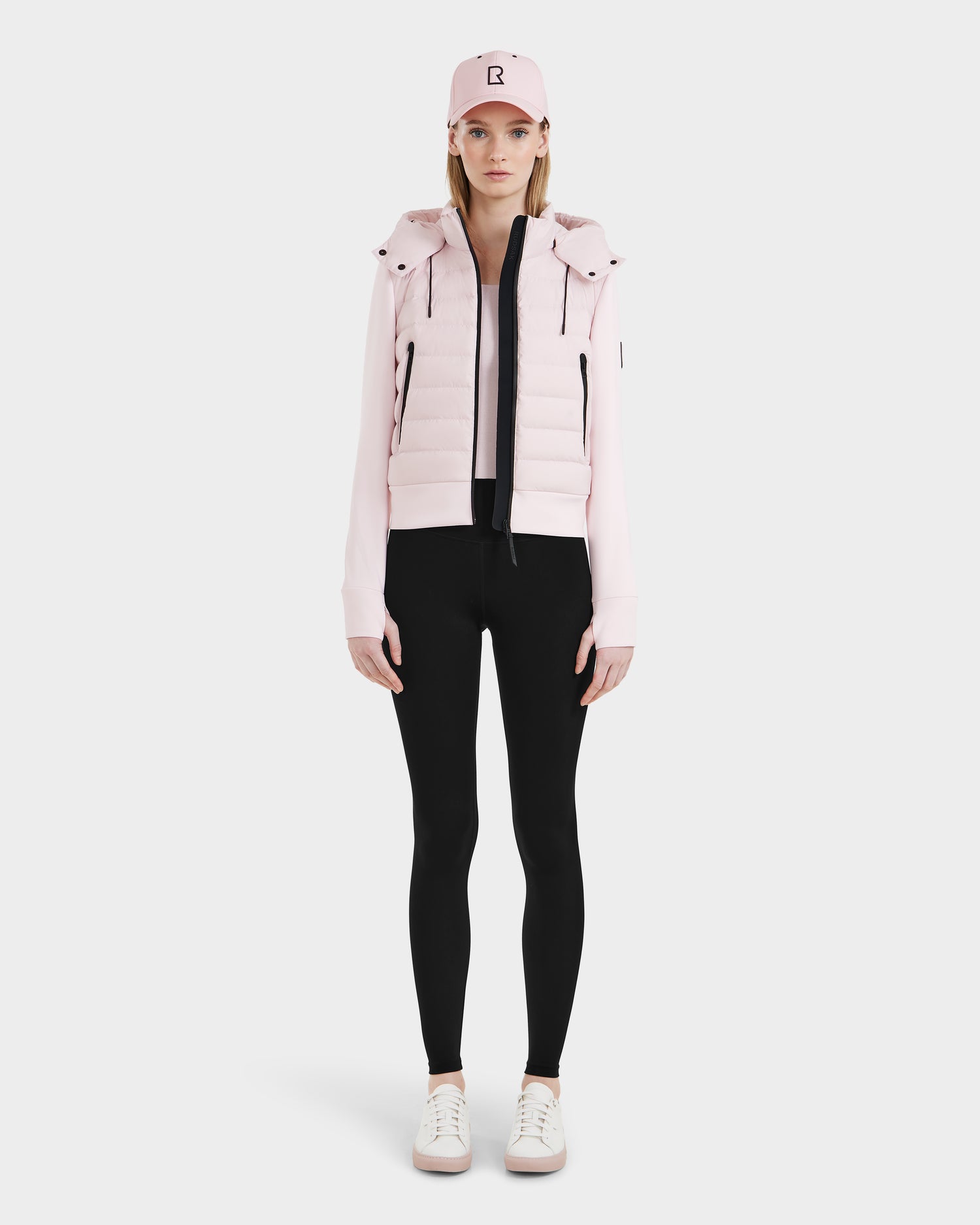 A woman stands facing forward against a white background wearing the Rudsak CASSA - 8125057 light pink cropped jacket, black leggings, white sneakers, and a matching light pink baseball cap with a B.